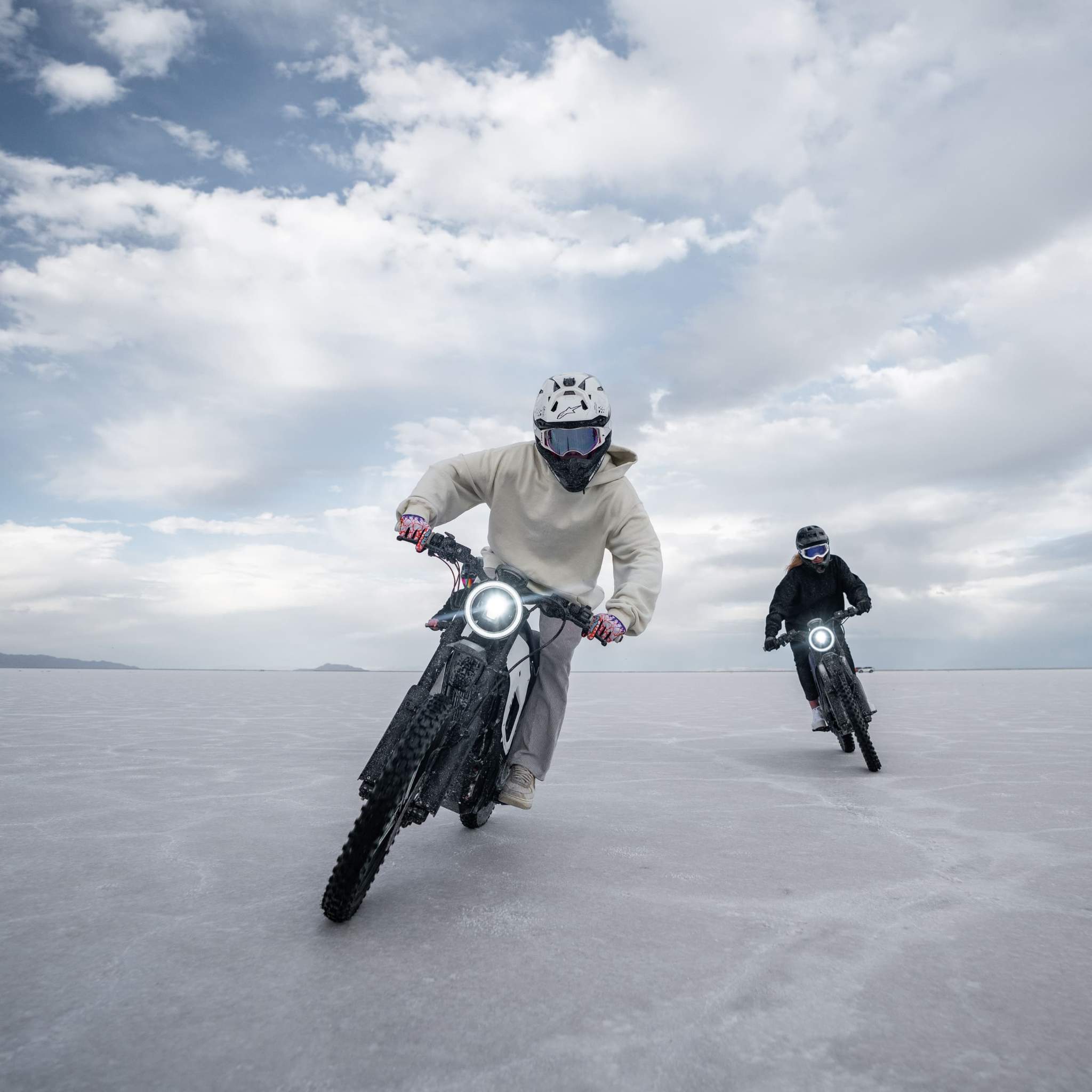 Zwei Fahrer mit Helmen und Ausrüstung fahren auf NIU XQi3 Enduro Dirtbike Cross Motorrad 45 km/h von NIU durch eine flache weiße Landschaft unter bewölktem Himmel, die Scheinwerfer leuchten hell, wobei der führende Fahrer näher an der Kamera steht.