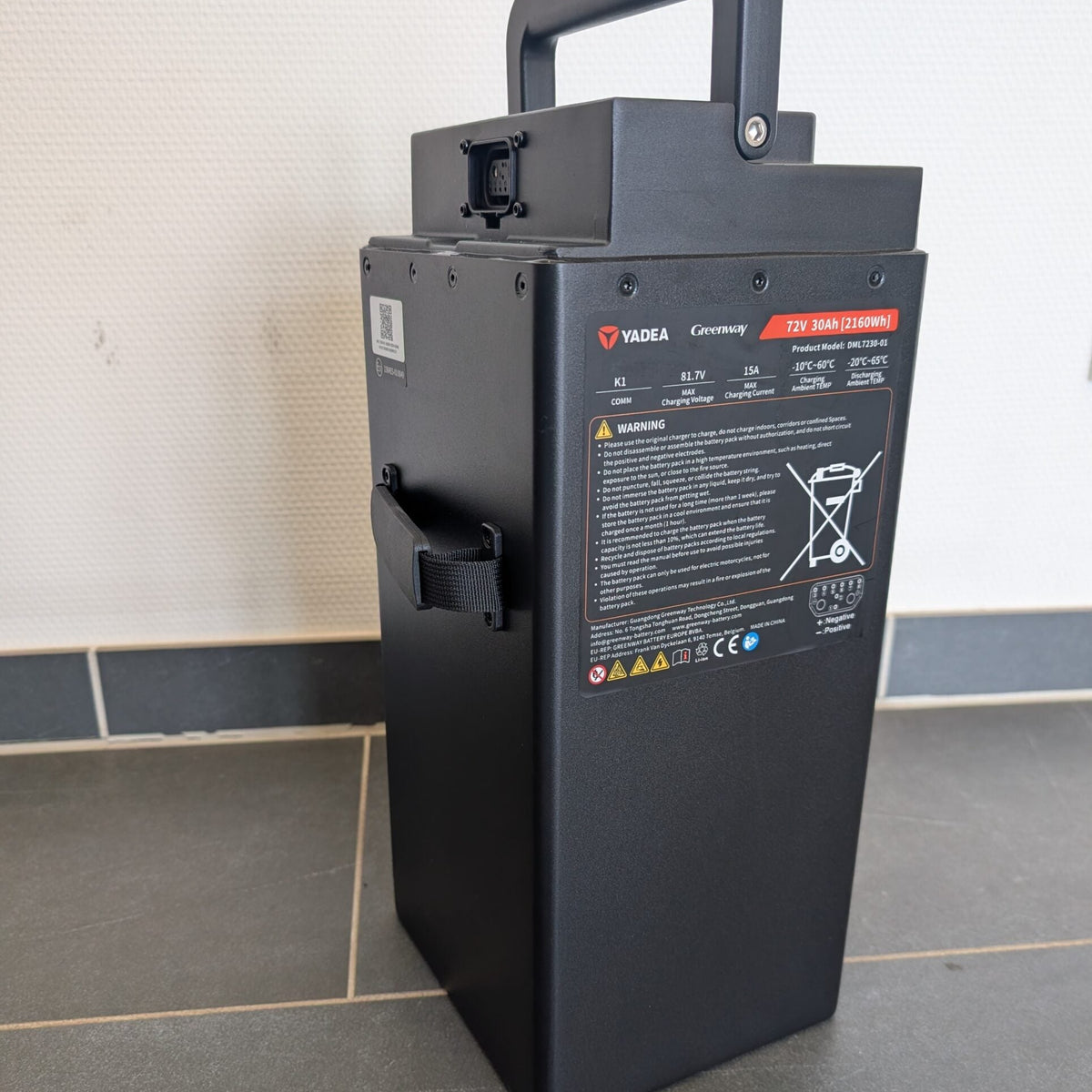 Ein schwarzer, rechteckiger Yadea-Lithium-LFP-Akku mit einem Griff steht auf einem Fliesenboden vor einer weißen Wand. An der Seite sind Warnaufkleber und technische Angaben zu sehen, die darauf hinweisen, dass er für den Einsatz im Yadea Owin U 2800W Retro-Elektroroller (45 km/h) geeignet ist.