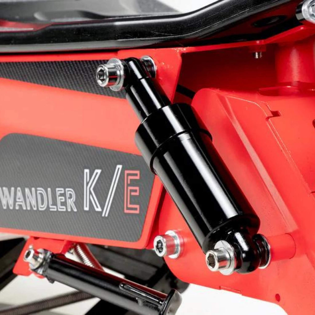 Nahaufnahme eines schwarzen Stoßdämpfers, der an einem rot-schwarzen mechanischen Rahmen montiert ist; an der Seite ist teilweise der Aufdruck „WANDLER K/E“ zu erkennen – ein Beleg für die robuste Technik des Wandler K/E 45 km/h E-Rollers / E-Scooter mit Sitz und Straßenzulassung, der für den Einsatz als Elektrofahrzeug im städtischen Verkehr konzipiert ist.