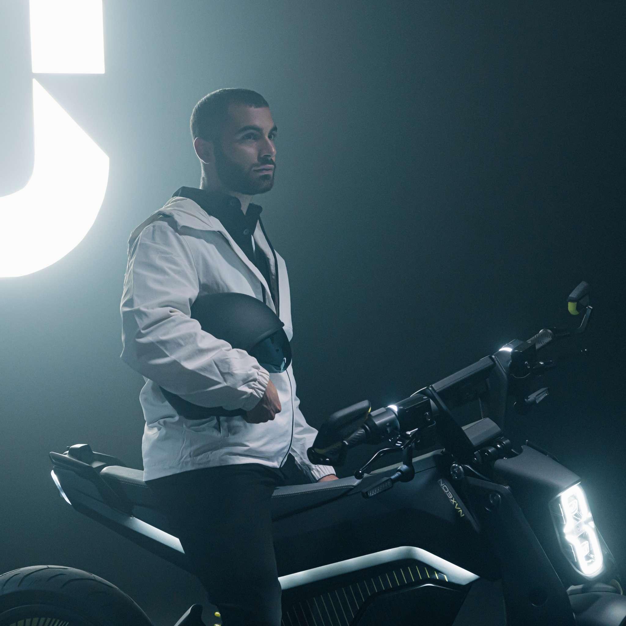 Ein Mann in einer weißen Jacke hält einen schwarzen Helm neben dem schnittigen NAXEON E-Motorrad Naxeon I AM. PRO (Kopie) in einem schwach beleuchteten Raum, wobei die Turbo-Boost-Funktionen durch helle Lichter im Hintergrund beleuchtet werden.
