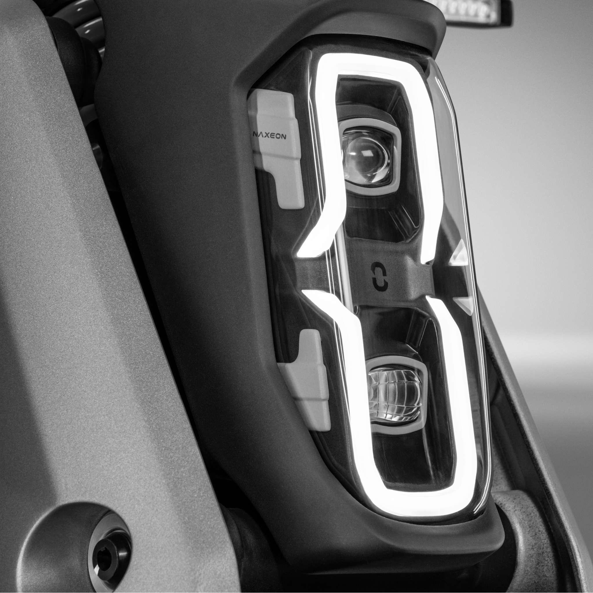 Nahaufnahme, schwarz-weiß Bild des NAXEON E-Motorrad Naxeon I AM. PRO (Copy) Scheinwerfer mit geometrischem LED-Tagfahrlicht um zwei Hauptglühbirnen, das das vom Turbo Boost inspirierte Design unterstreicht.