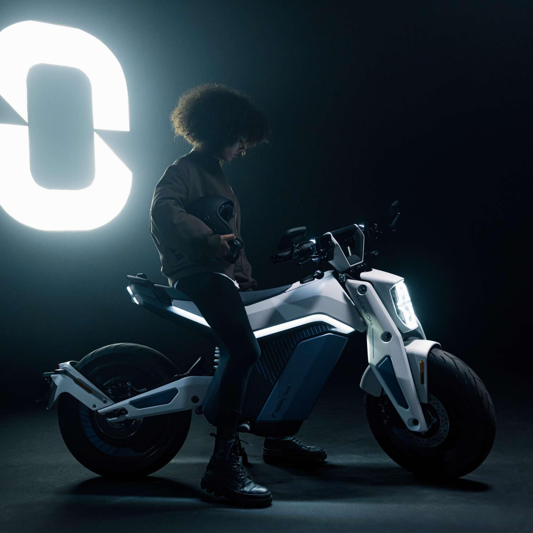 Eine Person mit lockigem Haar sitzt auf einem modernen weißen E-Motorrad Naxeon I AM. PRO von NAXEON in einem dunklen Raum, beleuchtet von einer großen leuchtenden Null im Hintergrund.
