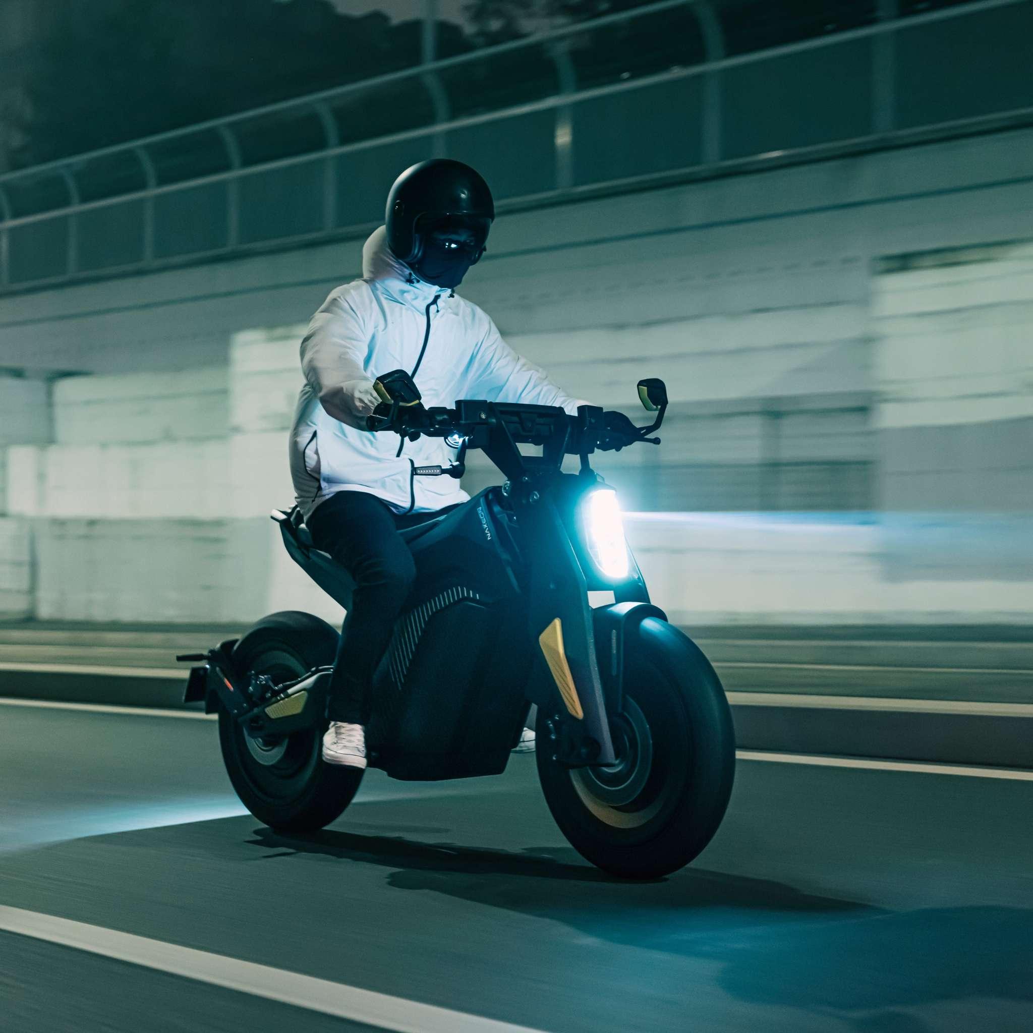 Eine Person in weißer Jacke und schwarzem Helm fährt auf dem NAXEON E-Motorrad Naxeon I AM. PRO (Copy) Elektromotorrad durch eine nächtliche Stadtstraße, mit unscharfen Lichtern, die im Hintergrund leuchten.