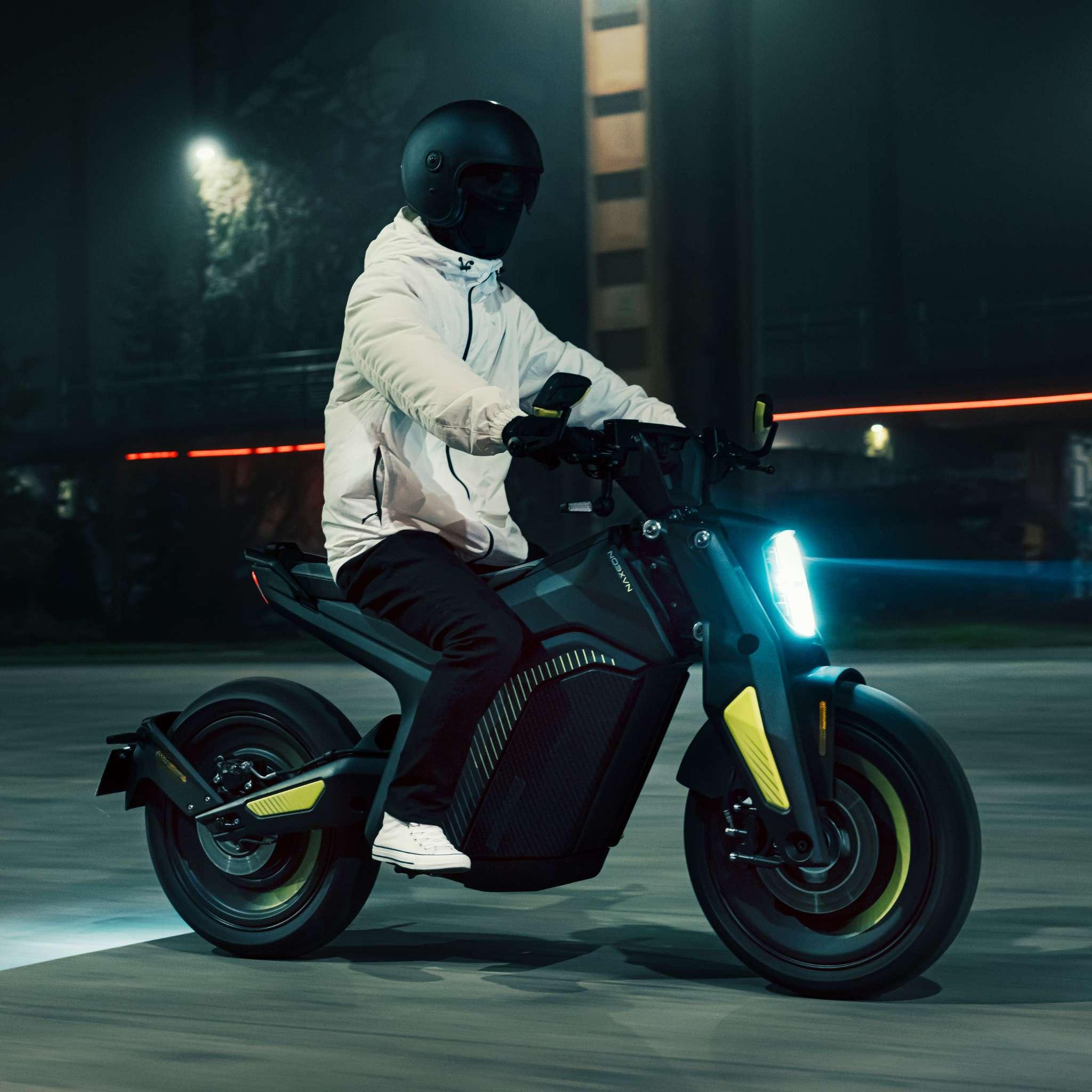 Eine Person in weißer Jacke, schwarzer Hose und schwarzem Helm fährt mit dem NAXEON E-Motorrad Naxeon I AM. PRO (Copy) bei Nacht auf einer Straße in der Stadt, beleuchtet von seinem hellen Scheinwerfer.