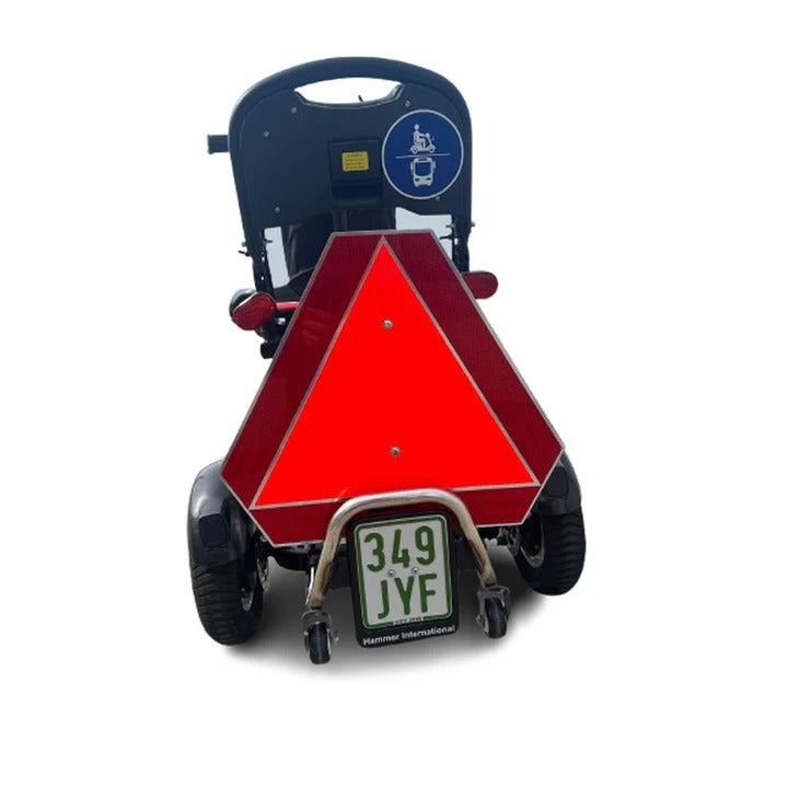 Das SONS OF ELECTRICITY Mobot Krankenfahrstuhl / Seniorenmobil 15 km/h - faltbar, leicht & mit Straßenzulassung verfügt über ein großes rotes Langsamfahrstellenschild, ein grün-weißes Nummernschild und einen blauen Rollstuhlaufkleber auf dem Sitz.