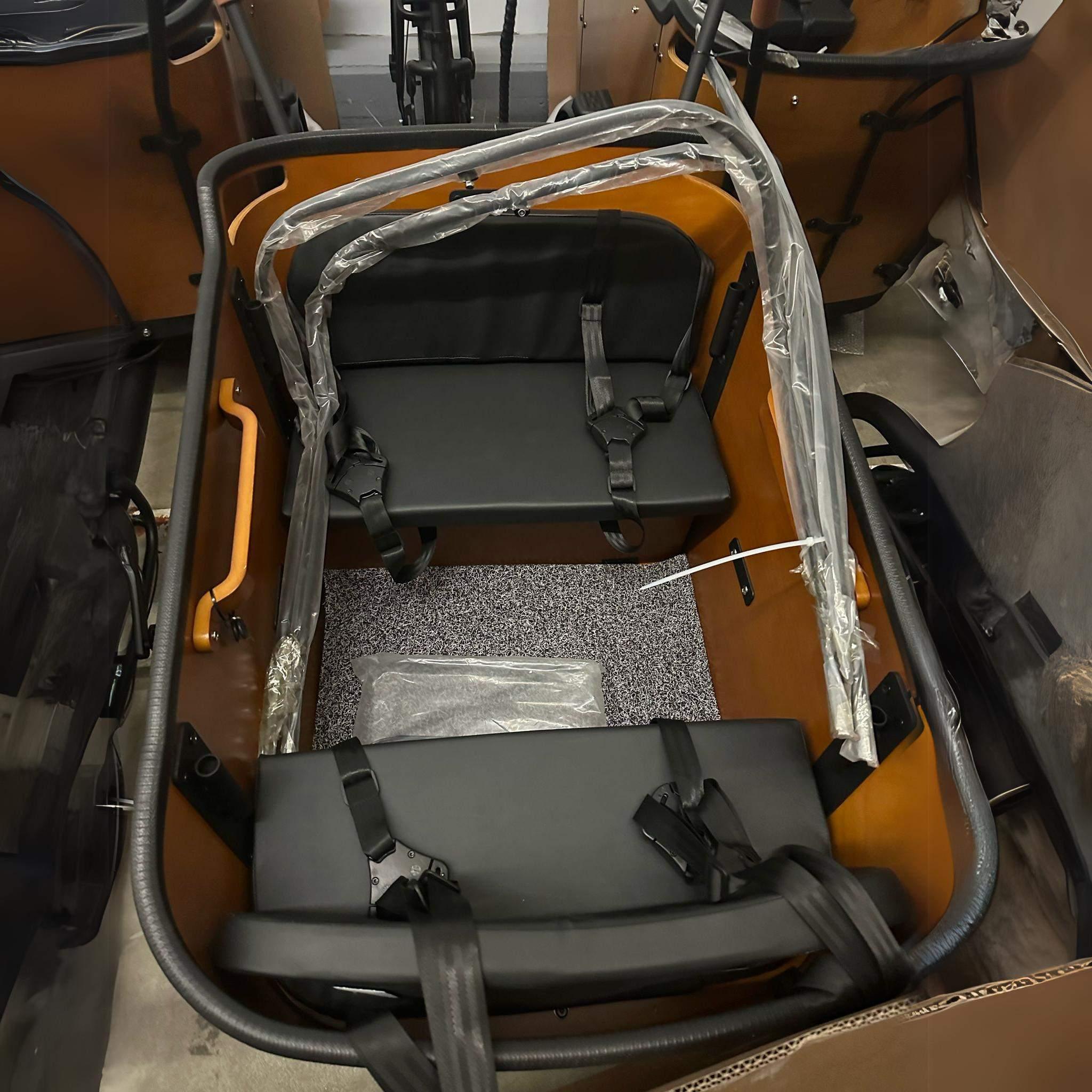 Draufsicht auf das leere Abteil des Tinbot Elektro Lastenrad TB-B1/B2: zwei schwarze gepolsterte Sitzbänke mit Gurten und Schutzhülle, braune Innenausstattung und eine gesprenkelte Bodenmatte. Marke: Tinbot, inklusive Regenschutz.