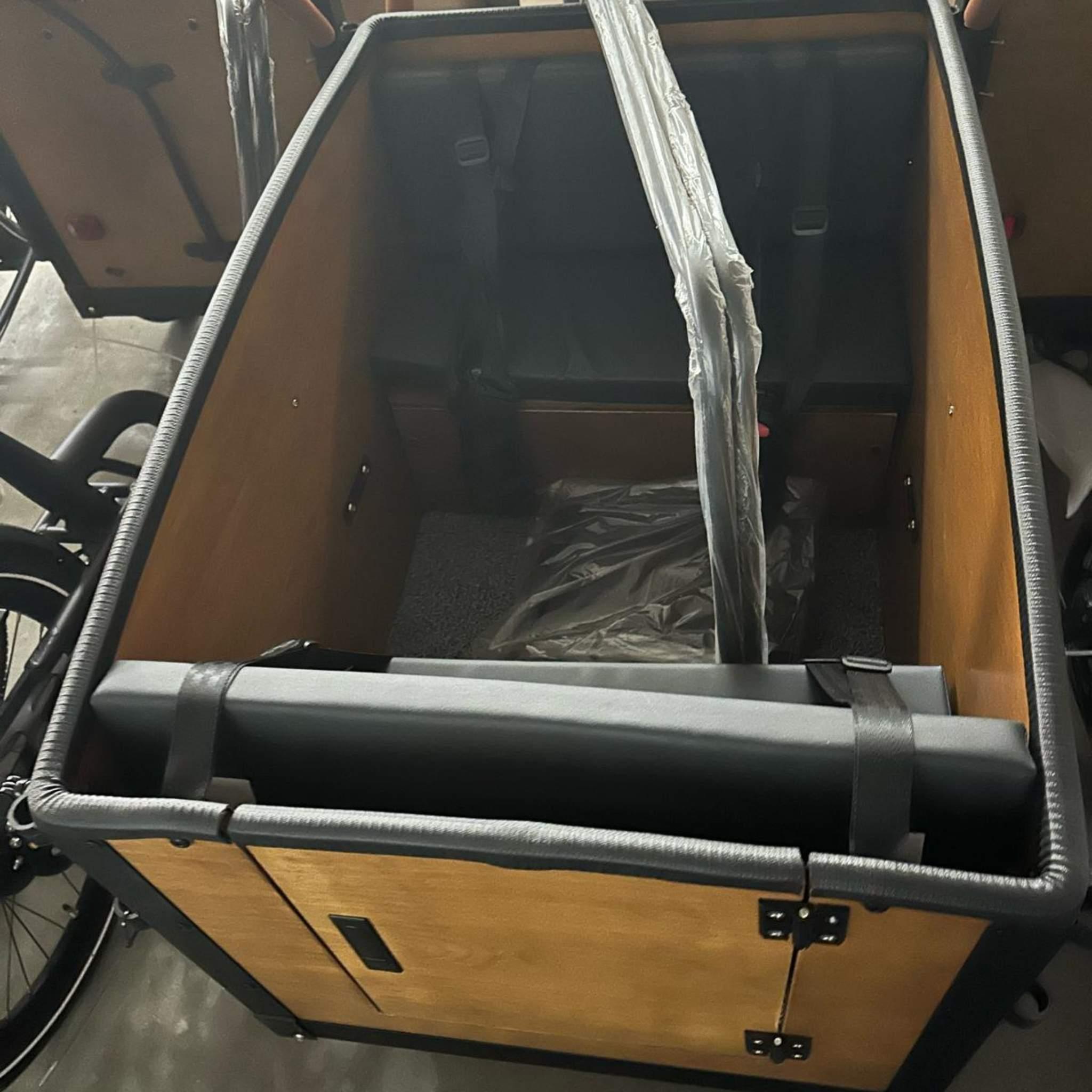 Eine Nahaufnahme der leeren hölzernen Ladekiste auf einem Tinbot Elektro Lastenrad TB-B1/B2 zeigt gepolsterte Sitze, Sicherheitsgurte, kunststoffummantelte Kissen, Metallscharniere und einen schwarzen gepolsterten Rand; einschließlich Regendach.