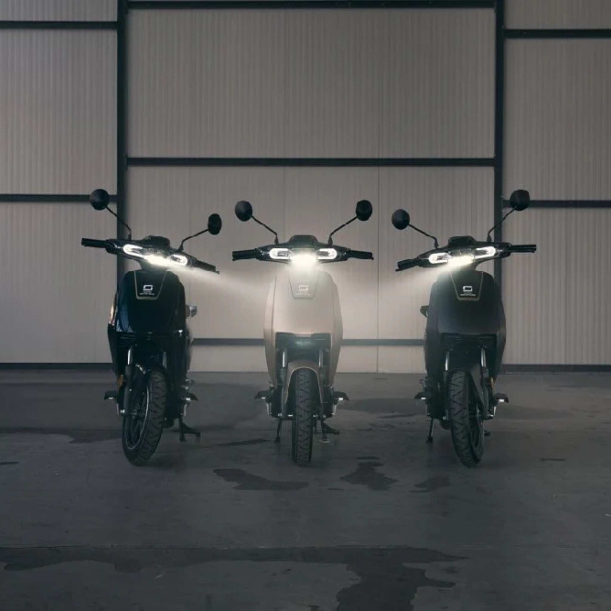 Drei E-Scooter-Modelle, darunter der Super Soco Elektroroller CUx Pro (45 km/h, Bosch-Power), stehen mit beleuchteten Scheinwerfern nebeneinander in einem großzügigen, schwach beleuchteten Innenraum vor einer metallverkleideten Wand.