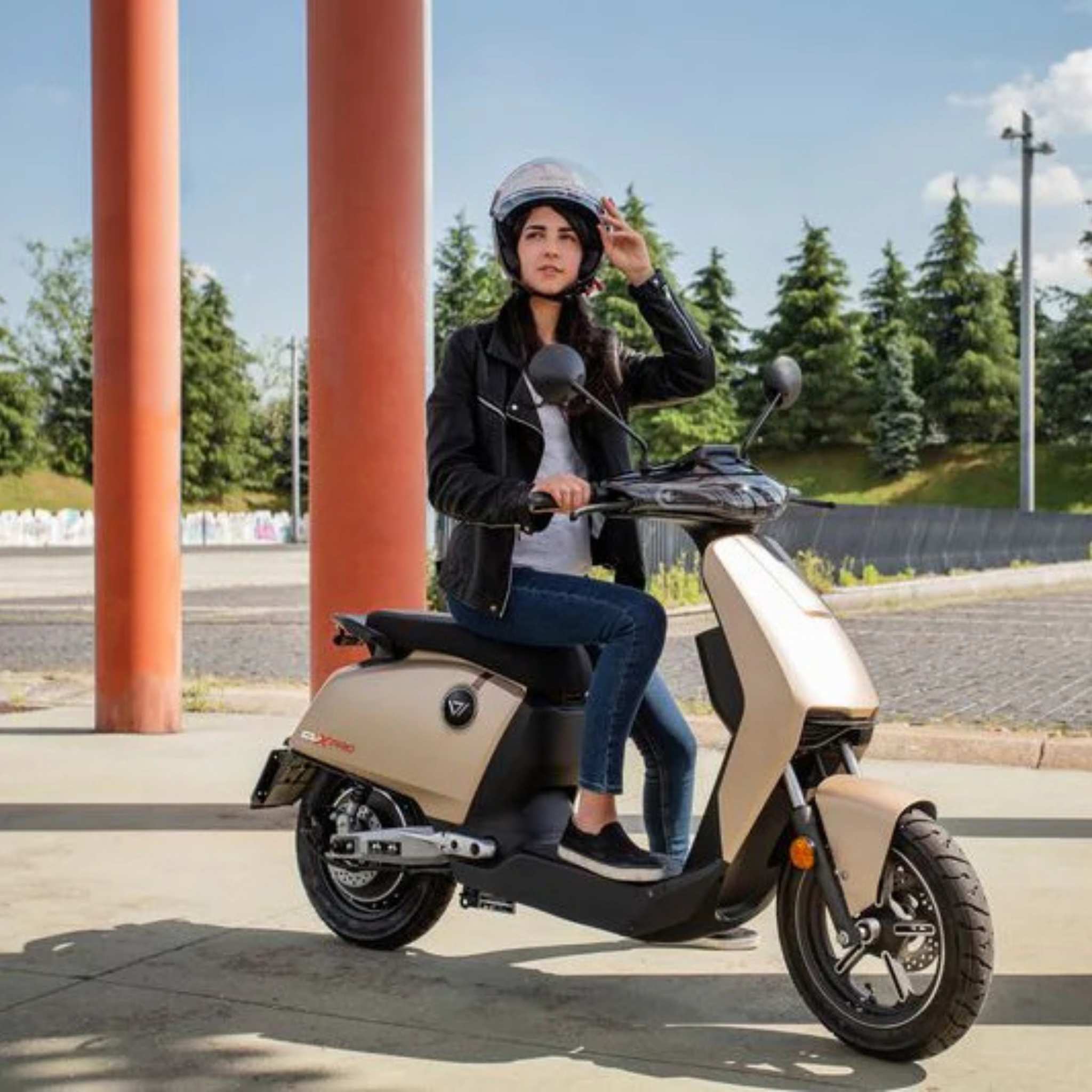 Eine Frau mit Helm und schwarzer Lederjacke sitzt im Freien auf einem beigen Super Soco Elektroroller CUx Pro, dem leichten 45 km/h E-Roller mit Bosch-Power, in der Nähe hoher roter Säulen und Bäume vor einem klaren Himmel.