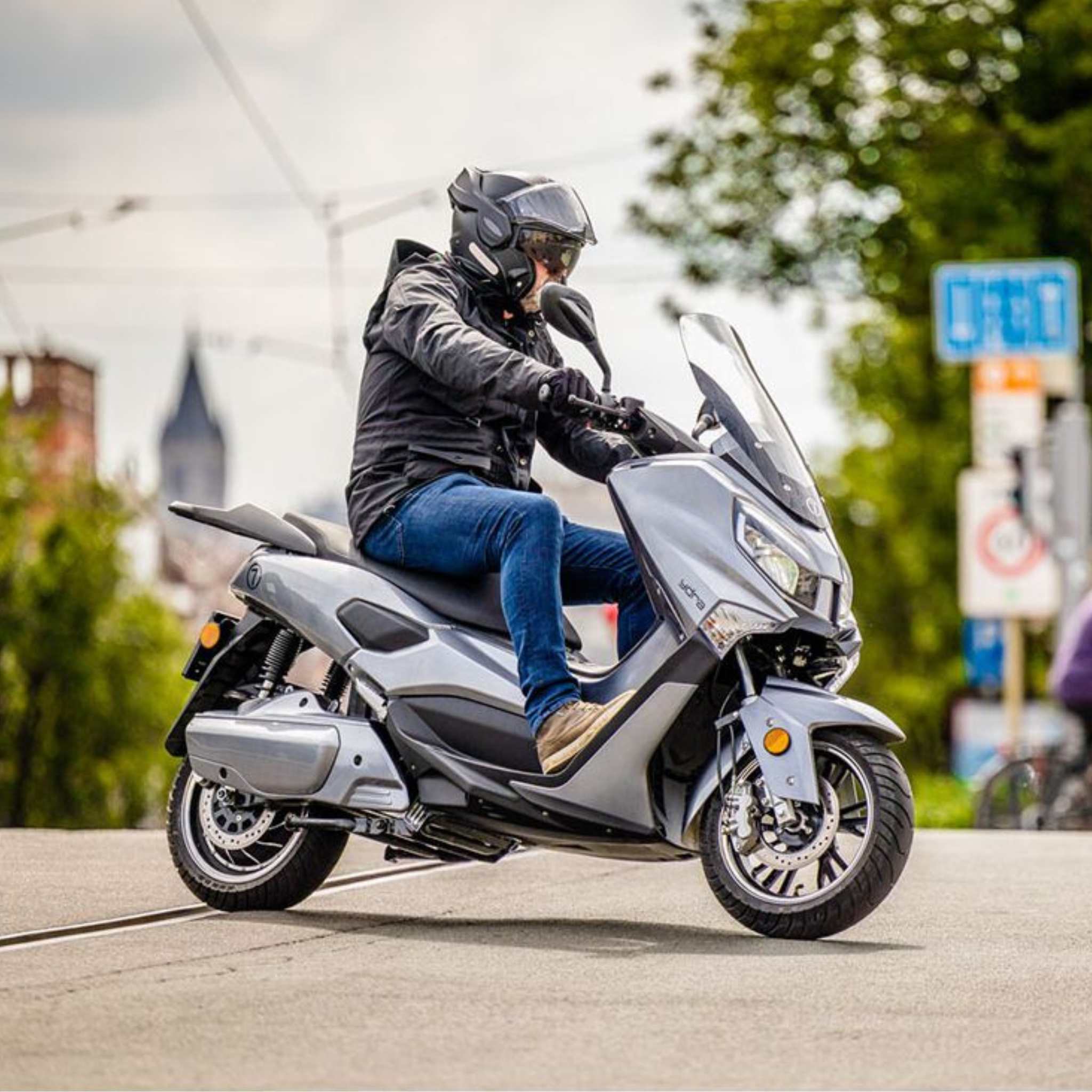 Eine Person mit Helm, Jacke, Jeans und Stiefeln fährt auf einem Trinity Jupiter GT 300 Elektroroller (120 km/h, 300 km Reichweite) eine Stadtstraße entlang, die von grünen Bäumen und unscharfen Gebäuden im Hintergrund gesäumt ist.