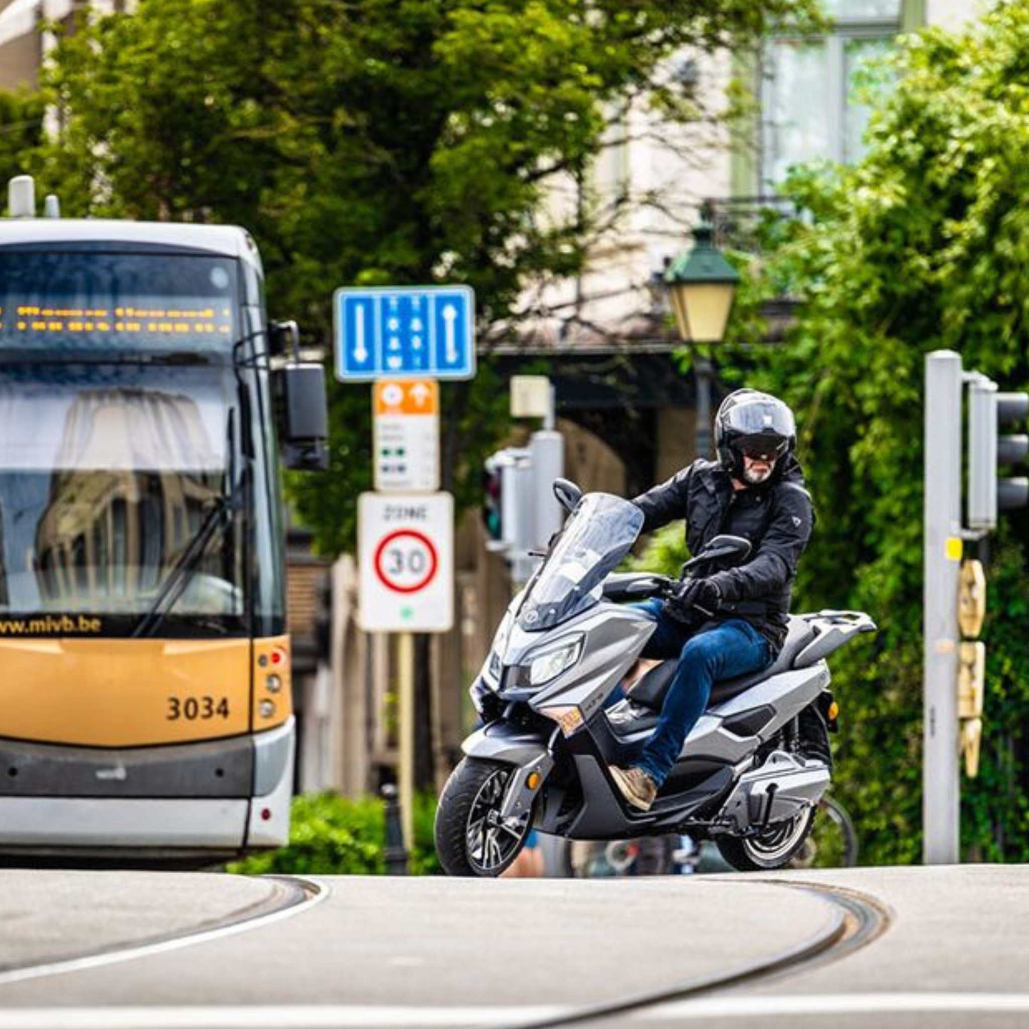 Ein behelmter Fahrer cruist auf dem Trinity Jupiter GT 300, einem Elektroroller von Trinity mit einer Reichweite von bis zu 300 km und einer Geschwindigkeit von 120 km/h, über eine belebte Straße in der Nähe einer gelben Straßenbahn - ideal für urbane Abenteuer zwischen grünen Bäumen und Verkehrszeichen.