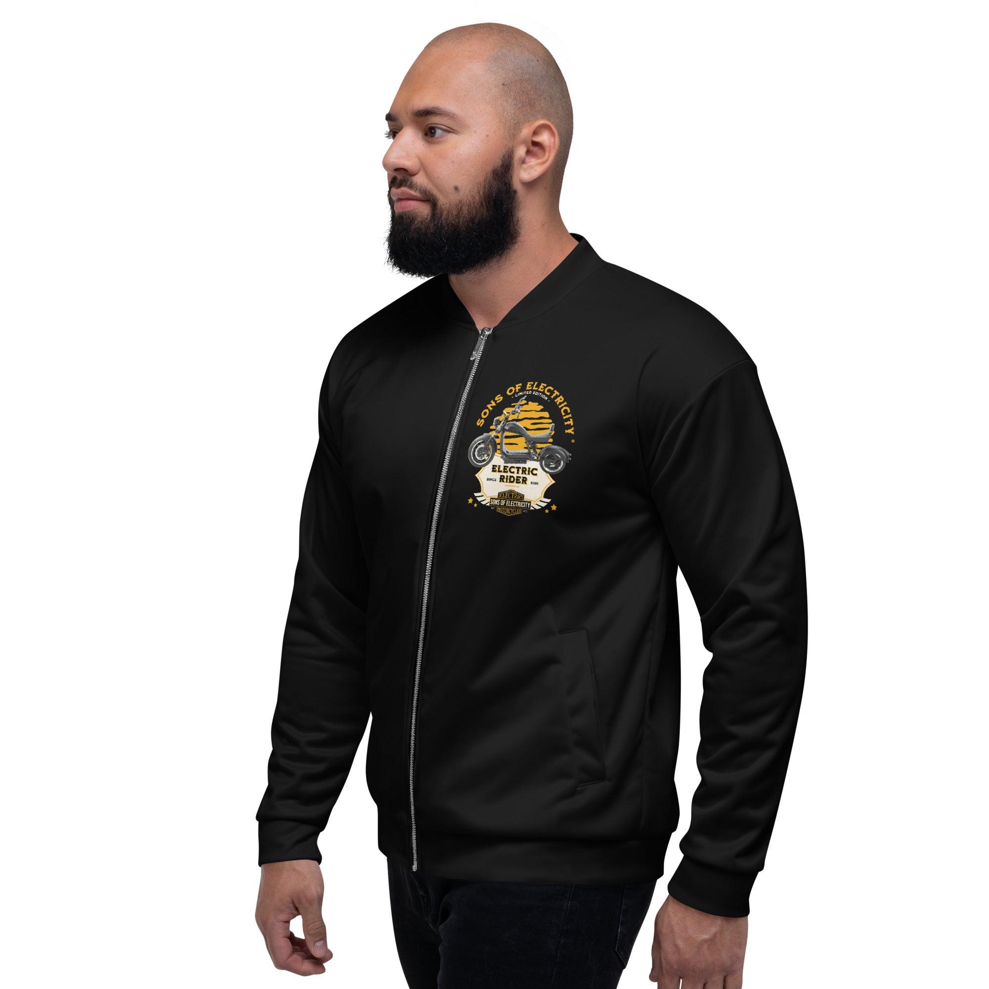 Ein bärtiger Mann steht seitlich und trägt eine schwarze SONS OF ELECTRICITY Premium E-Chopper (2) Bomberjacke Electric Rider Limited Edition, die ein Board of Electricity zeigt: Electrical Engineering Grafik mit Zahnrädern und Sternen auf der linken Brust.