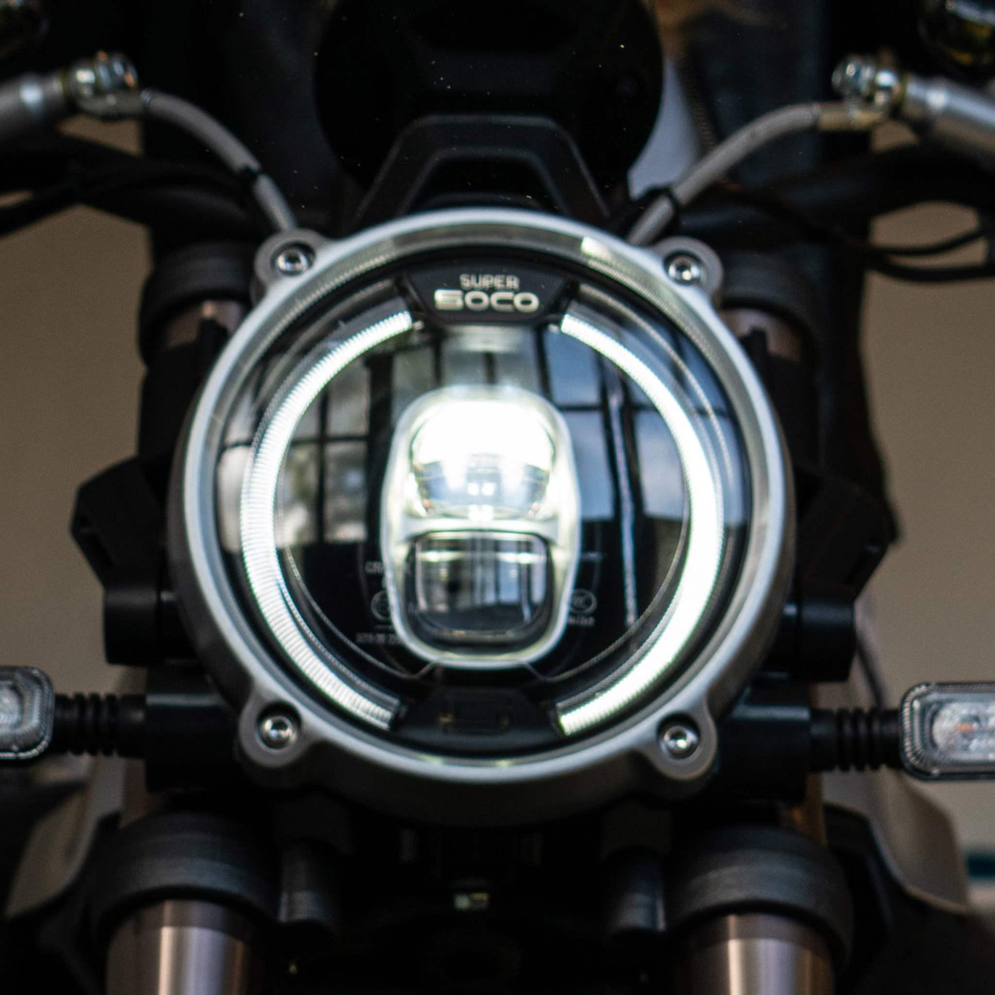 Nahaufnahme eines runden Motorradscheinwerfers mit einem hellen LED-Ring, beschriftet mit Super Soco, montiert auf dem E-Motorrad SUPER SOCO TC MAX 95 km/h; im Hintergrund sind Lenker und Verkabelung zu sehen.