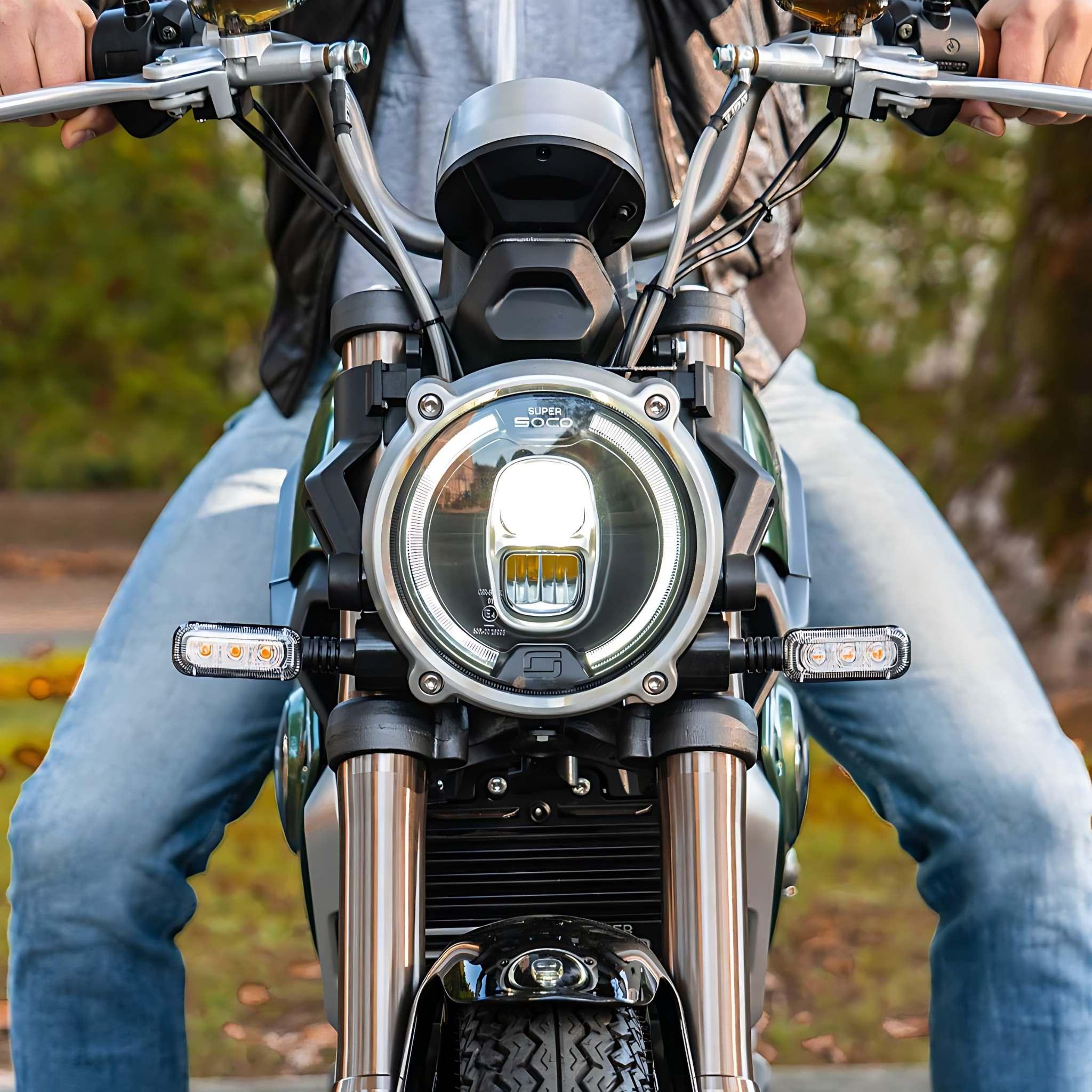 Eine Person in Lederjacke und Jeans sitzt auf einem Super Soco E-Motorrad SUPER SOCO TC Eco 45 km/h, gezeigt von vorne mit sichtbarem Scheinwerfer und Lenker, vor einem Hintergrund aus verschwommenem Grün.