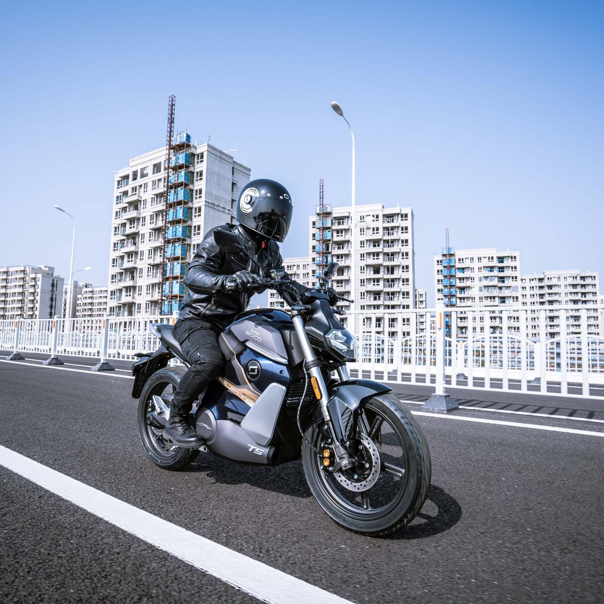 Ein Fahrer in schwarzer Motorradkleidung und mit Helm fährt mit dem Super Soco E-Motorrad TS Streethunter (75 km/h) auf einer leeren Stadtstraße, mit modernen Wohnhäusern und einem klaren blauen Himmel im Hintergrund.