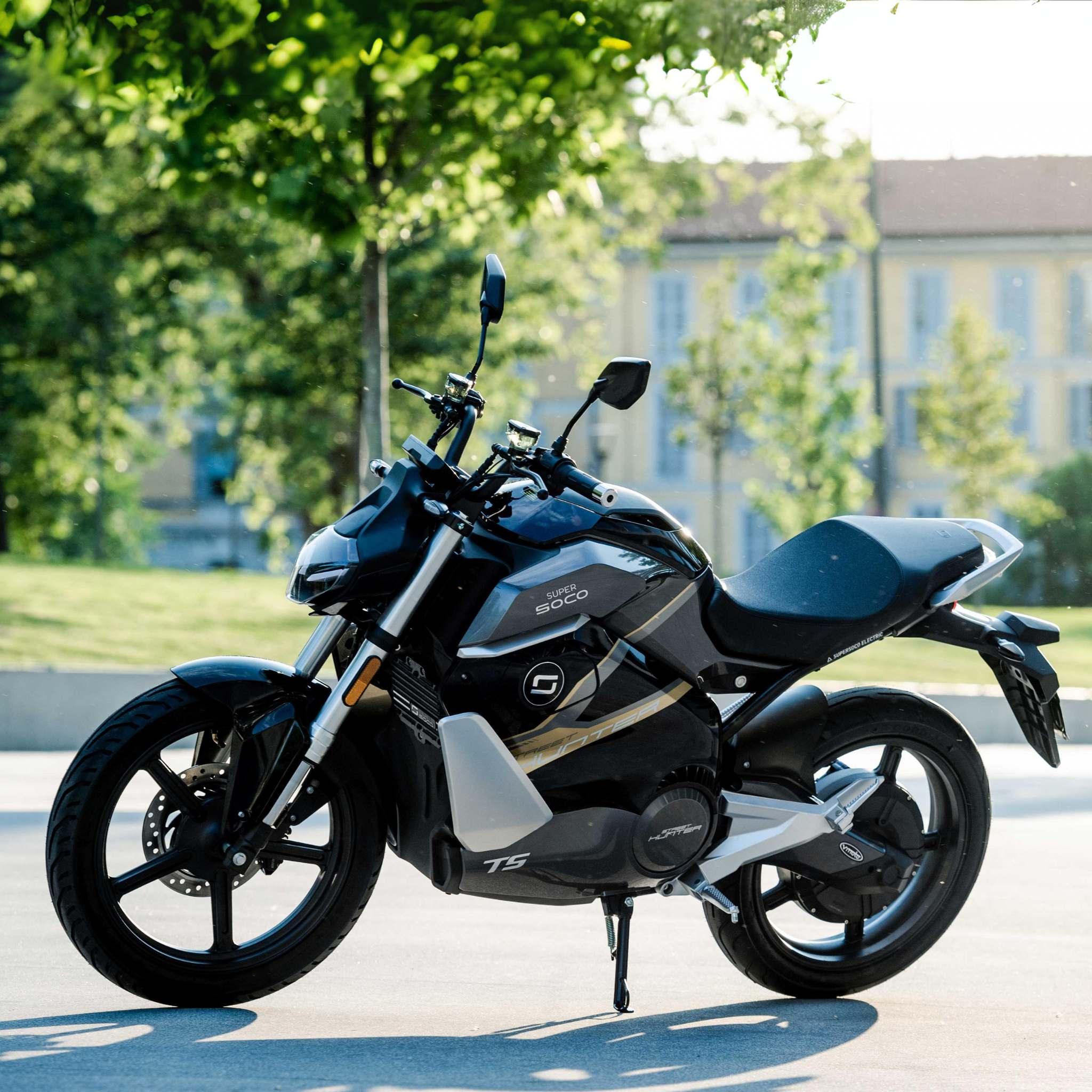 Ein Super Soco E-Motorrad TS Streethunter in Schwarz und Silber ist an einem sonnigen Tag im Freien geparkt, mit Bäumen und einem Gebäude mit großen Fenstern dahinter. Dieses Elektrofahrrad erreicht eine Geschwindigkeit von bis zu 75 km/h.