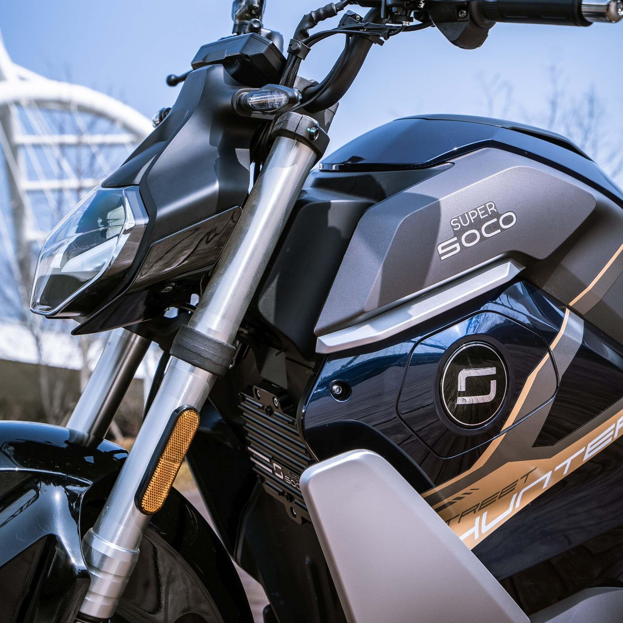 Nahaufnahme des E-Motorrads SUPER SOCO TS Streethunter 75 km/h von Super Soco, die den gebrandeten Tank, die Vorderradaufhängung und den geometrischen Scheinwerfer vor einem sanft verschwommenen städtischen Hintergrund zeigt. Höchstgeschwindigkeit: 75 km/h.