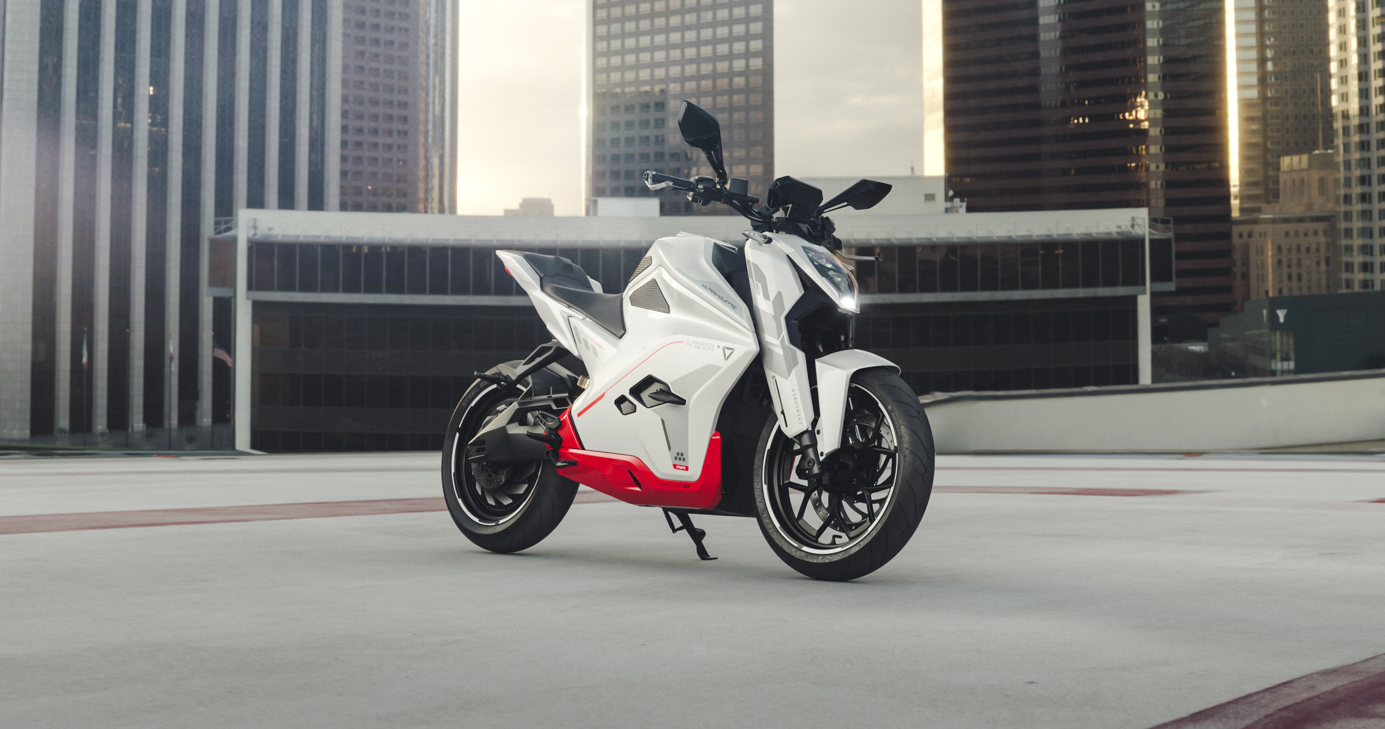 Ein Ultraviolette F77 Superstreet Recon - ein sportliches weiß-rotes Elektromotorrad von Ultraviolette - ist auf einem Dach geparkt, mit hohen modernen Gebäuden im Hintergrund unter einem bewölkten Himmel.