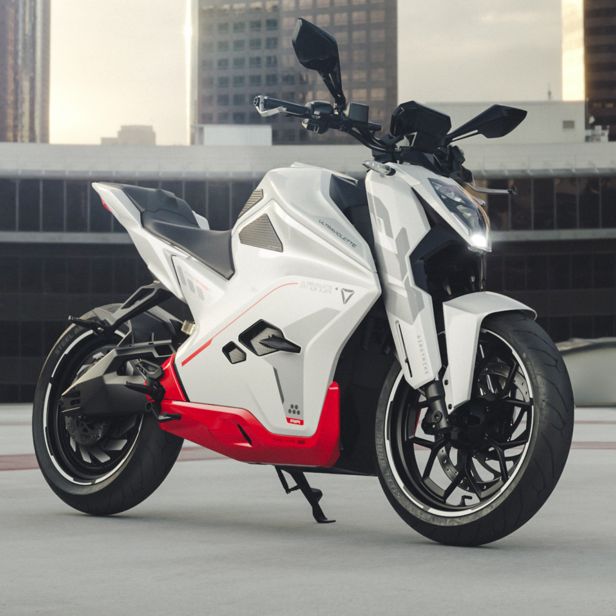 Ein Ultraviolette F77 Superstreet Recon - ein sportliches weiß-rotes Elektromotorrad von Ultraviolette - ist auf einem Dach geparkt, mit hohen modernen Gebäuden im Hintergrund unter einem bewölkten Himmel.