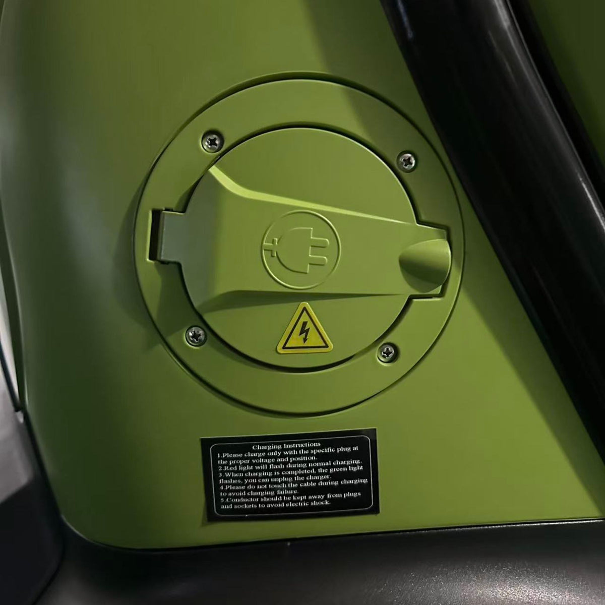 Nahaufnahme der grünen Abdeckung des Ladeanschlusses am City Twister Luqi EV300 Pro Kabinenroller (Modell 2026) mit einem Stecksymbol, einem Hochspannungswarnhinweis und einem kleinen Hinweisschild darunter.