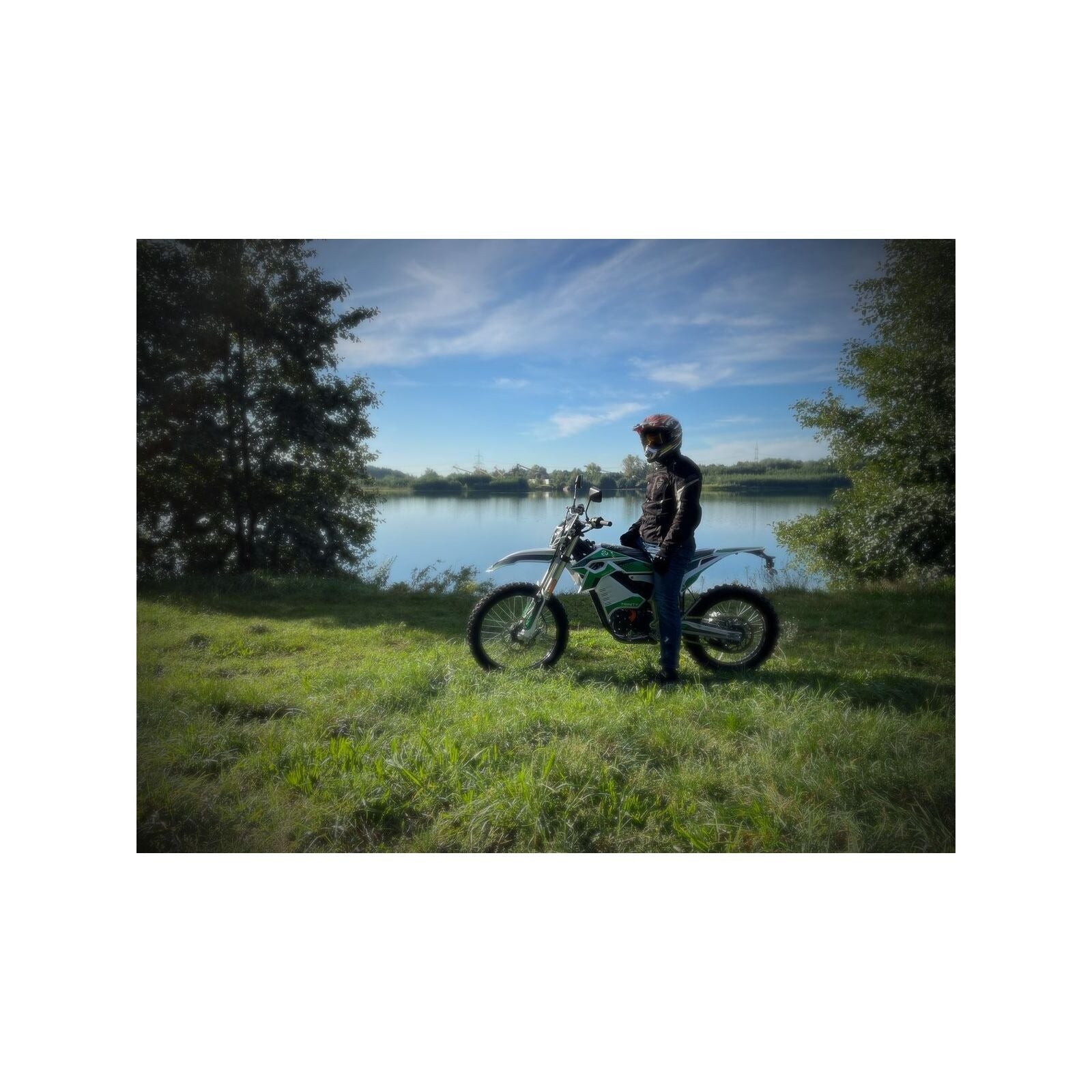 Eine Person mit Helm und Jacke steht neben einem Trinity Panthera II Enduro Elektromotorrad (1143 Nm, Schaltgetriebe, 125er Klasse) auf einer Wiese in der Nähe eines Sees, mit Bäumen und blauem Himmel im Hintergrund.