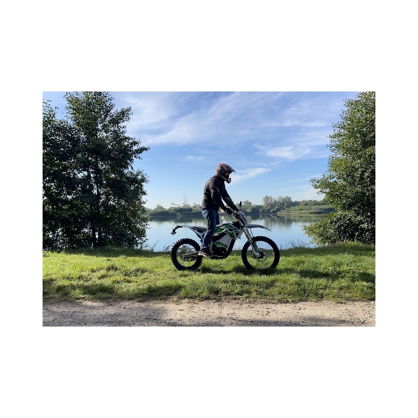 Ein Fahrer mit Helm und Jacke sitzt auf einem Trinity Panthera II Enduro Elektromotorrad (1143 Nm Drehmoment, Schaltgetriebe), geparkt auf einer Wiese an einem ruhigen See, mit Bäumen und blauem Himmel im Hintergrund.