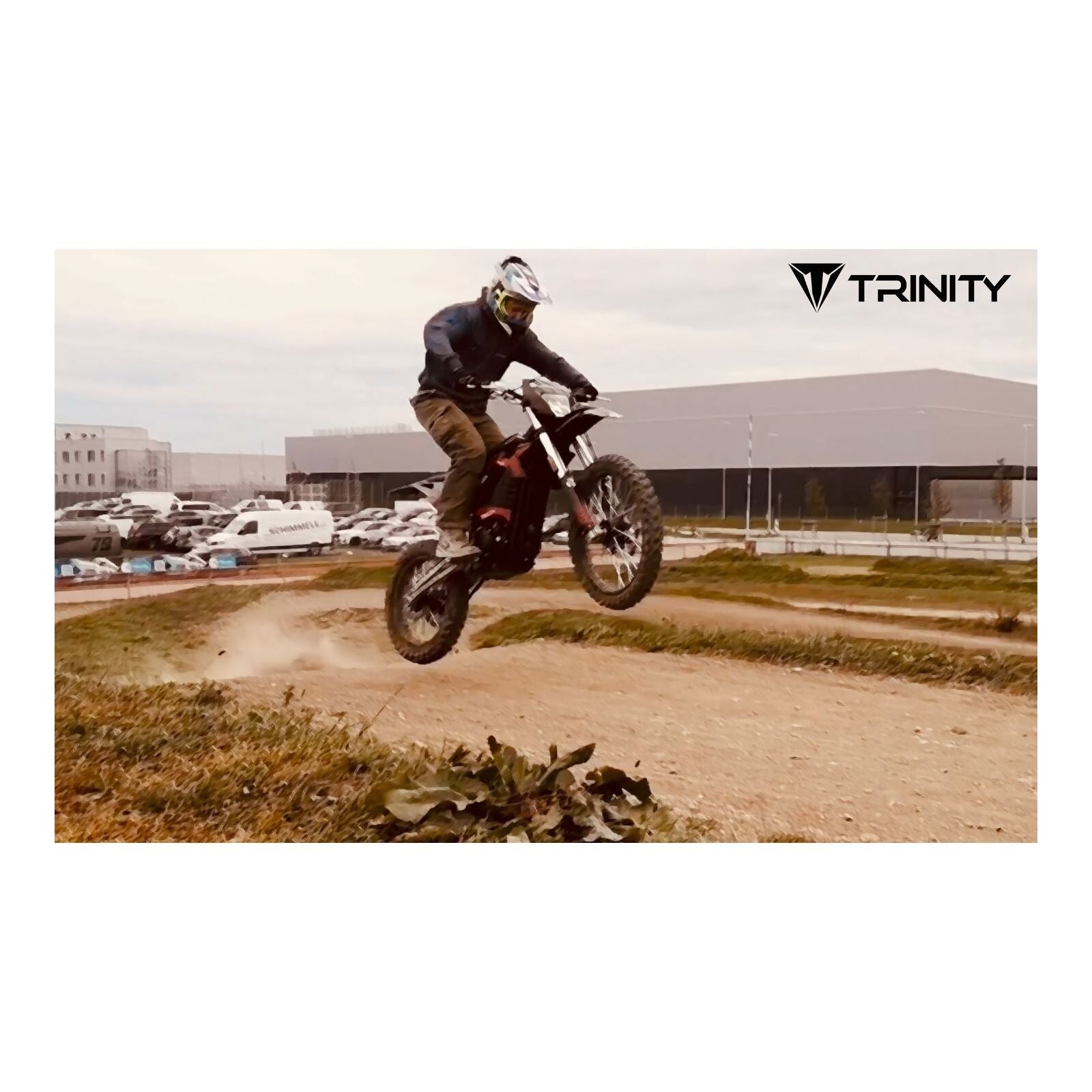 Ein Fahrer mit Helm lenkt ein Trinity Panthera II Enduro Elektromotorrad (1143 Nm Drehmoment, Schaltgetriebe) über einen Dirt-Track-Sprung, im Hintergrund sind das TRINITY-Logo, Gebäude und geparkte Autos zu sehen.