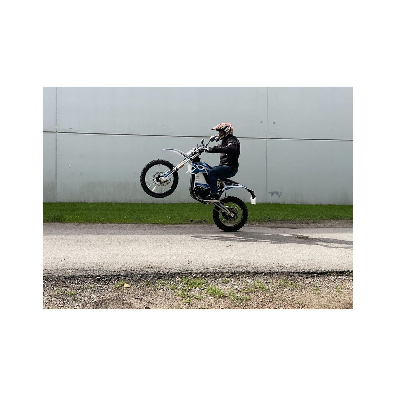 Ein behelmter Fahrer in einer schwarzen Jacke vollführt einen Wheelie auf einem Trinity Panthera II Enduro Elektromotorrad (1143 Nm Drehmoment, Schaltgetriebe) auf dem Bürgersteig vor einer großen grauen Wand.