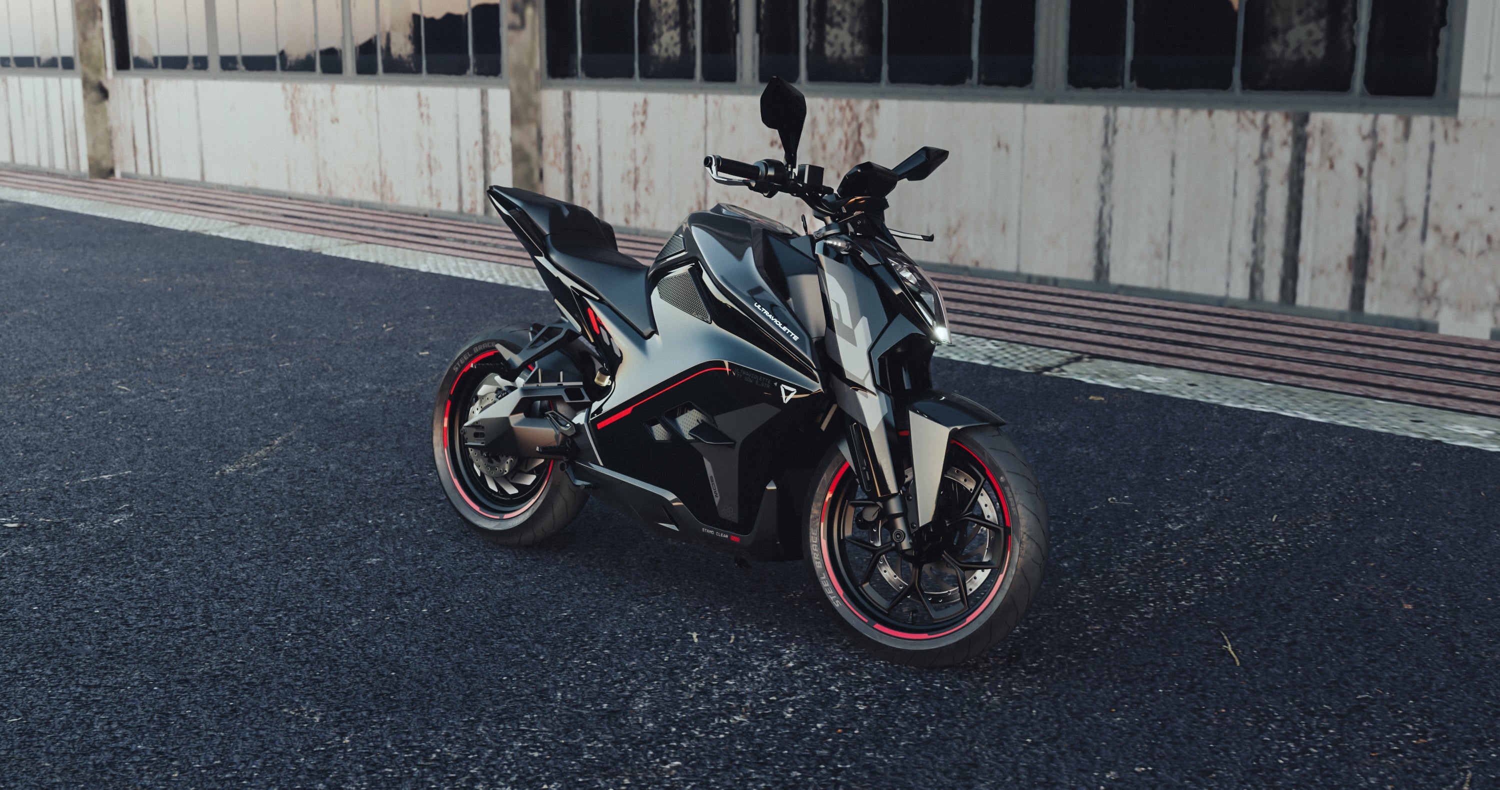 Die Ultraviolette F77 Superstreet Recon, ein schnittiges schwarz-silbernes Elektromotorrad mit roten Akzenten, ist auf dunklem Asphalt vor einem verwitterten Gebäude mit großen Fenstern geparkt.