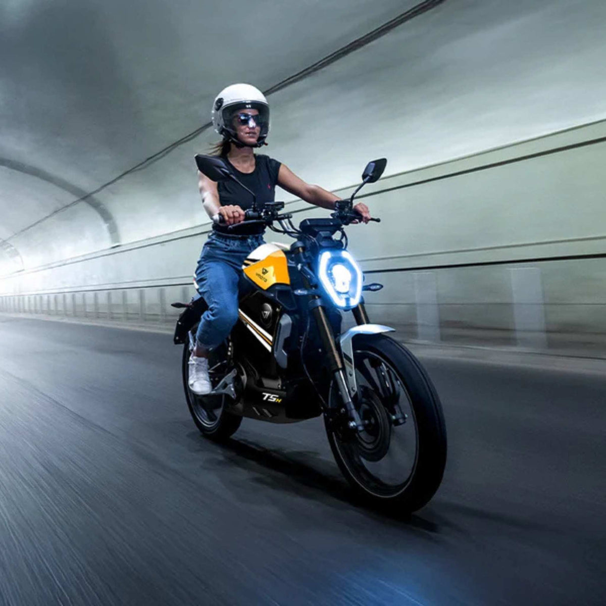 Ein Fahrer mit Helm, Sonnenbrille und Freizeitkleidung rast auf dem VMOTO TSX 72V, einem leistungsstarken 45 km/h schnellen Elektroroller im markanten Motorrad-Design von VMOTO, durch einen gut beleuchteten Tunnel, wobei Bewegungsunschärfen die schnelle Fahrt unterstreichen.