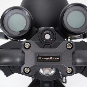 Nahaufnahme des Armaturenbretts des Mangosteen M1P Citycoco E-Chopper (MODELL 26) mit zwei runden Anzeigen, einem Zündschalter mit der Aufschrift „IGNITION“ und einem Schlüsselschlitz sowie einer Lenkerklemme mit Mangosteen-Logo. Dieser E-Chopper wird von einem robusten 2000-Watt-Elektromotor angetrieben und ist mit einem zuverlässigen 30-Ah-Lithium-Ionen-Akku ausgestattet, der für verbesserte Leistung und Geschwindigkeiten von bis zu 45 km/h sorgt.