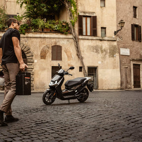 Ein Mann mit einer schwarzen Schachtel überquert eine Kopfsteinpflasterstraße und geht auf einen geparkten NIU Elektroroller E-Scooter FQI 500 (2025, 95 km/h) zu, der vor einem alten, beigen Gebäude mit Bogenfenstern und Weinreben steht und moderne elektrische Mobilität widerspiegelt.