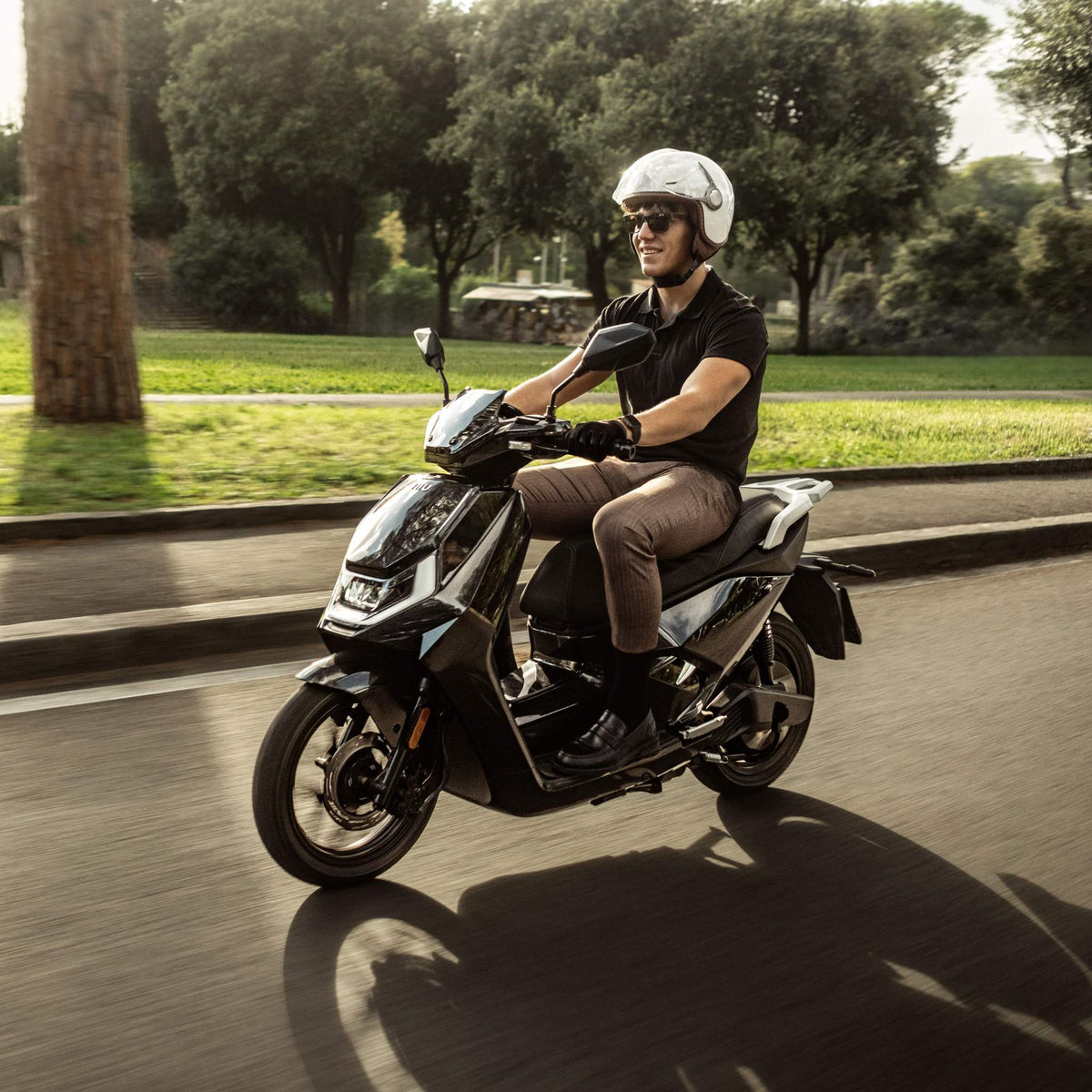 Ein Mann mit weißem Helm und Sonnenbrille fährt mit dem NIU Elektroroller E-Scooter Motorroller FQI 500 (95 km/h, 2025) eine sonnige, von Bäumen gesäumte Straße entlang und genießt die elektrische Mobilität.