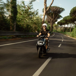 Ein behelmter Fahrer fährt auf einem schwarzen NIU Elektroroller E-Scooter Motorroller FQI 500 (2025, 95 km/h) eine von Bäumen gesäumte, kurvenreiche Straße entlang und genießt an einem sonnigen Tag die elektrische Mobilität.