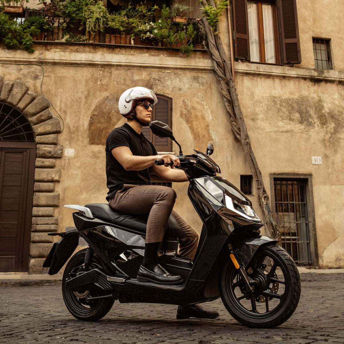 Ein Mann mit weißem Helm, Sonnenbrille, schwarzem T-Shirt und brauner Hose fährt mit einem NIU Elektroroller E-Scooter Motorroller FQI 500 (95 km/h, 2025) auf einer Kopfsteinpflasterstraße an einem alten Gebäude mit verschlossenen Fenstern und üppiger Bepflanzung vorbei.