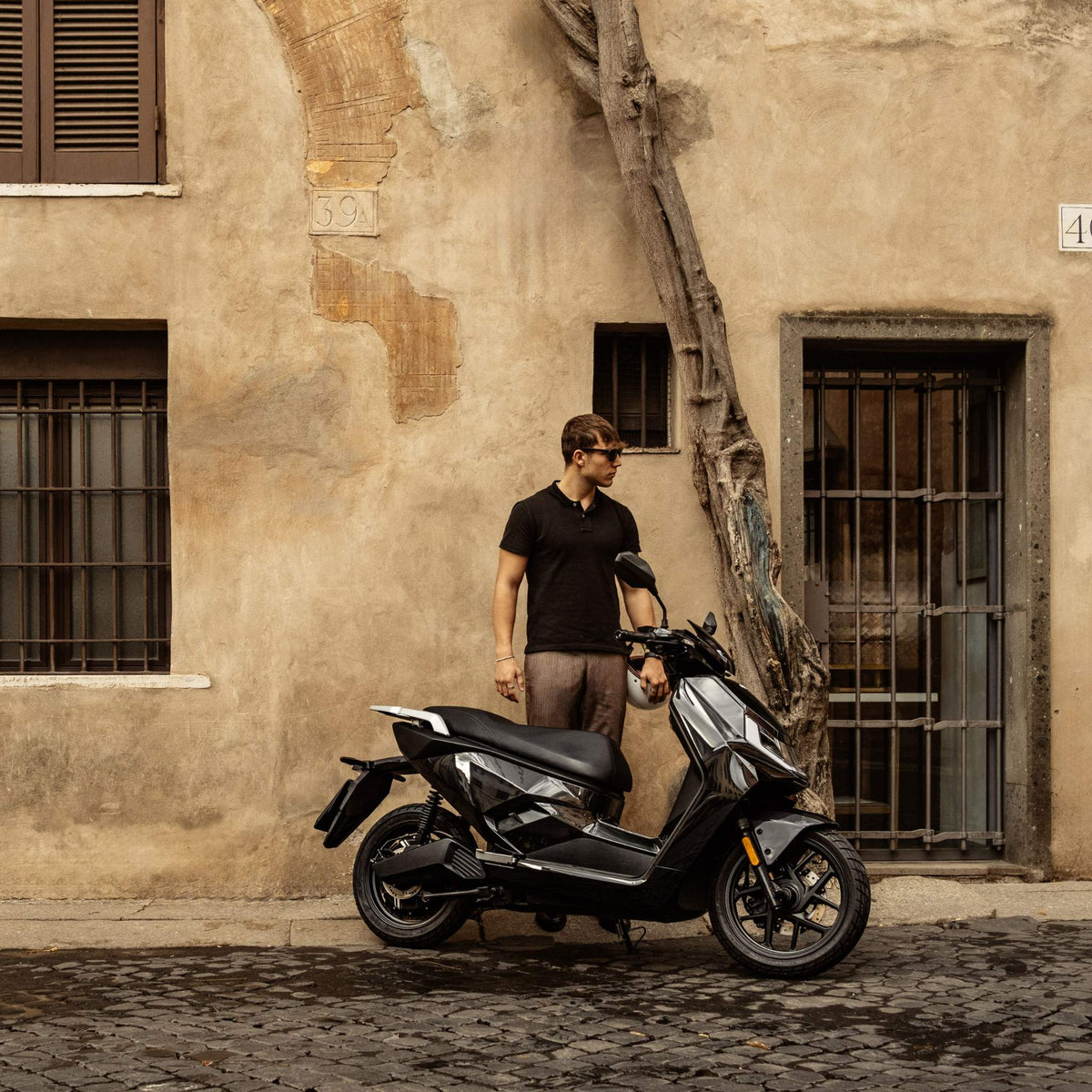 Ein Mann in einem schwarzen Hemd und mit Sonnenbrille steht neben einem NIU Elektroroller E-Scooter Motorroller FQI 500 (95 km/h, 2025) auf einer Kopfsteinpflasterstraße vor einem alten beigen Gebäude mit verschlossenen Fenstern und einem Baum in der Nähe.