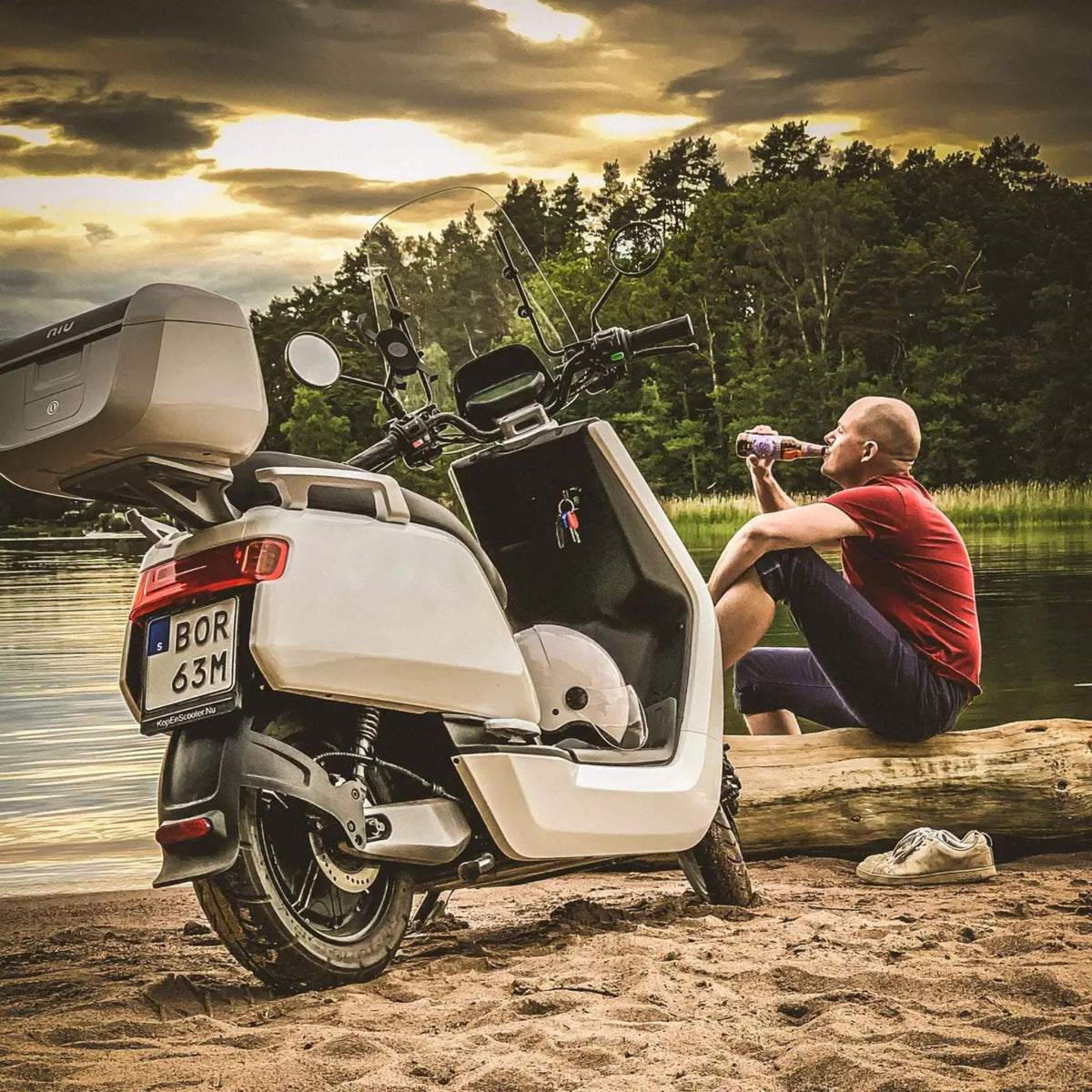Ein Mann im roten Hemd sitzt auf einem Baumstamm an einem See und trinkt aus einer Flasche. Neben ihm am sandigen Ufer steht der NIU Elektroroller Motorroller NQI Sport (2024 Upgrade), der die moderne Elektromobilität widerspiegelt. Bäume und Wolken vervollständigen die Szene.