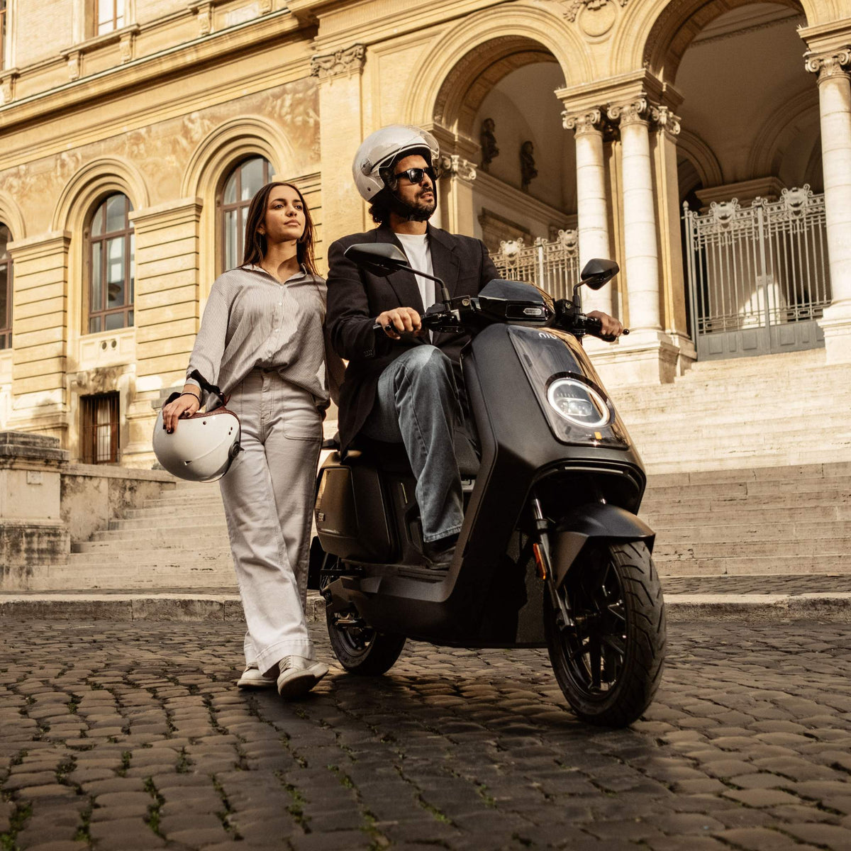 Ein Mann mit Helm sitzt auf einem schwarzen NIU NQiX 500 Elektroroller E-Scooter Motorroller Modell 2025 (100 km/h – SONDERPREIS!) von NIU, während eine Frau in Freizeitkleidung neben ihm steht und einen weißen Helm in der Hand hält; beide posieren vor einem beeindruckenden historischen Gebäude mit Steinstufen und Bögen.