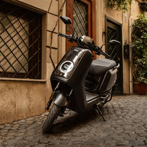 Ein schwarzer NIU NQiX 500 Elektroroller (E-Scooter) Modell 2025 der Marke NIU steht auf einer Kopfsteinpflasterstraße neben einem Gebäude mit vergitterten Fenstern und Grünpflanzen im Eingangsbereich.