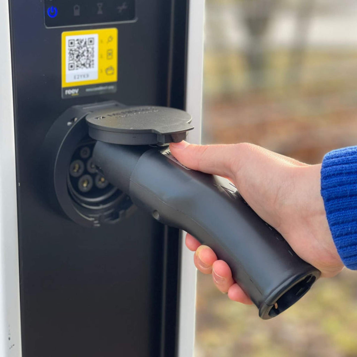 Eine Person im blauen Ärmel steckt den VMOTO TYP2 Adapter für Schuko Stecker in eine Outdoor-Ladestation mit QR-Code und blauem Power-Knopf, bereit zum Aufladen von Elektro Motorrad, E-Roller, E-Scooter und mehr.