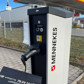 Ein VMOTO TYP2 Adapter für Schuko Stecker - die zuverlässige Ladelösung für dein Elektro Motorrad, E-Chopper, E-Roller & mehr - liegt an einer Ladesäule auf Pflaster nahe einer Tankstelle. Im Hintergrund sind Gras und ein Zaun zu sehen.