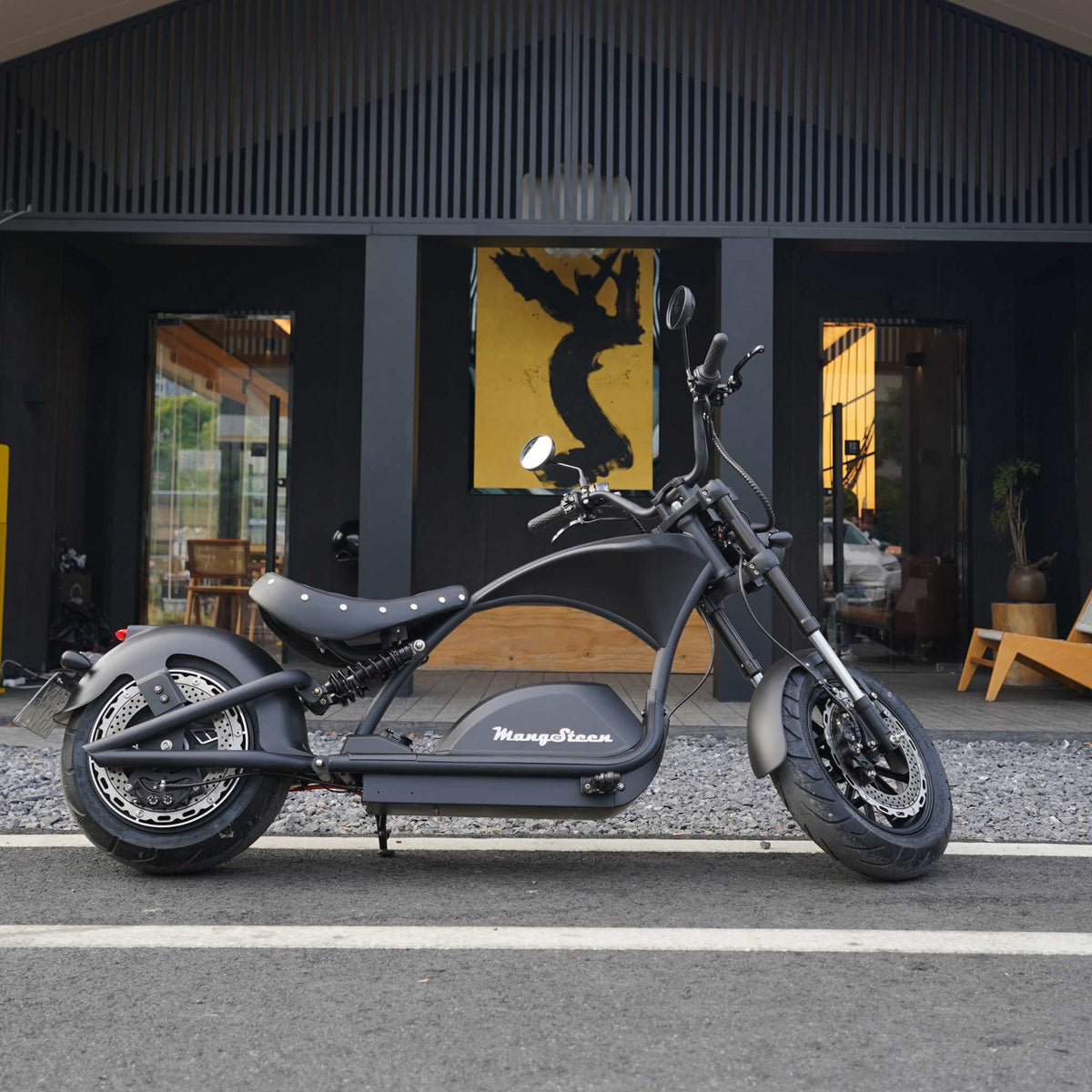 Ein stilvoller schwarzer Mangosteen E-Chopper (M1PS, 85 km/h, 40-Ah-Lithium-Akku von Samsung, 4000 Watt) steht auf einer Straße vor einem modernen Gebäude mit großen Fenstern, in dessen Inneren sich ein gelbes Kunstwerk befindet.