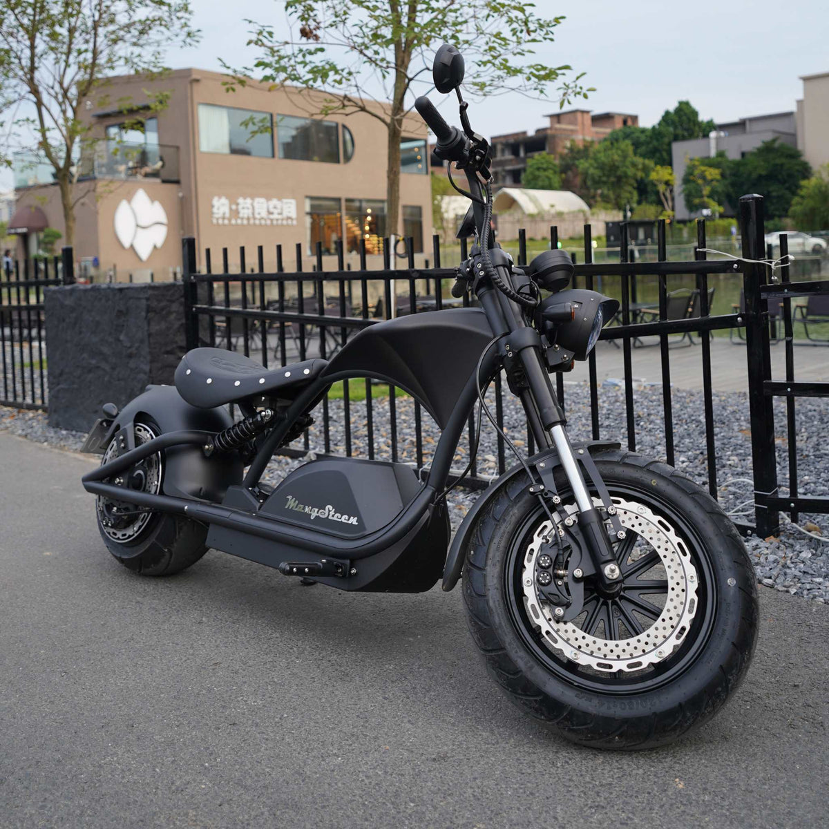 Ein mattschwarzes Mangosteen E-Chopper-Elektromotorrad, Modell M1PS, mit einem 4000-Watt-Motor und einer 40-Ah-Lithiumbatterie von Samsung steht auf einer Straße in der Nähe eines schwarzen Zauns, vor dem Hintergrund eines modernen Gebäudes und von Bäumen.