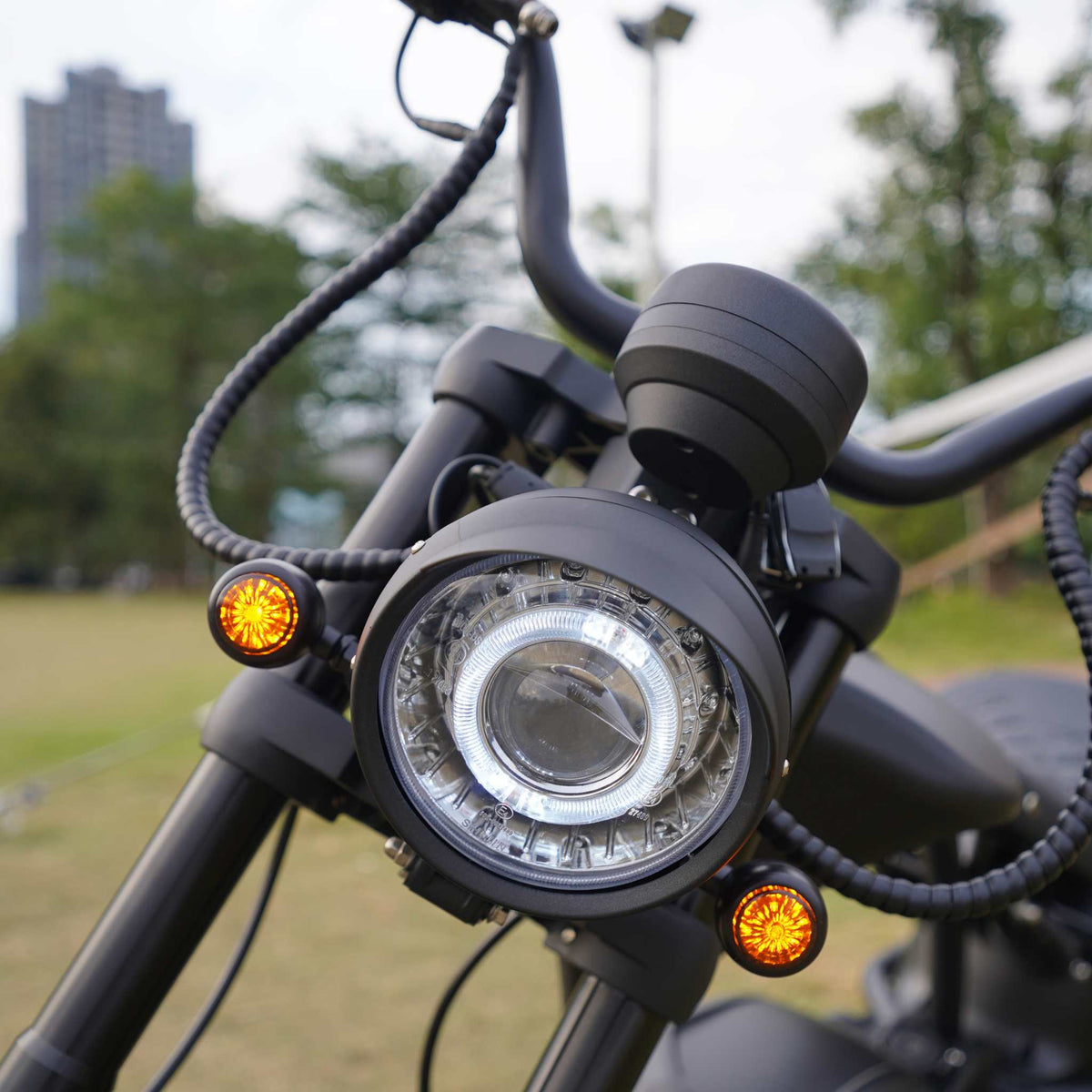 Nahaufnahme des Scheinwerfers und des Lenkers eines schwarzen Mangosteen Elektromotorrads M1PS (E-Chopper/E-Scooter, 4000 Watt, 85 km/h, 40 Ah Samsung-Lithium-Akku), mit zwei kleinen orangefarbenen Lichtern auf jeder Seite, im Freien auf einer Rasenfläche geparkt, im Hintergrund sind Bäume und ein Gebäude unscharf zu erkennen.