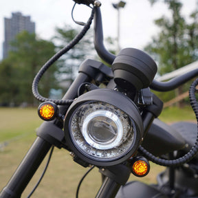 Nahaufnahme des Scheinwerfers und des Lenkers eines schwarzen Mangosteen Elektromotorrads M1PS (E-Chopper/E-Scooter, 4000 Watt, 85 km/h, 40 Ah Samsung-Lithium-Akku), mit zwei kleinen orangefarbenen Lichtern auf jeder Seite, im Freien auf einer Rasenfläche geparkt, im Hintergrund sind Bäume und ein Gebäude unscharf zu erkennen.