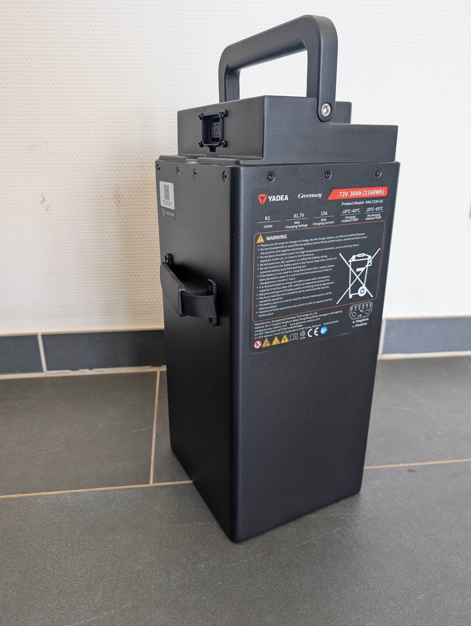 Ein schwarzer, rechteckiger Yadea-Lithium-LFP-Akku mit einem Griff steht auf einem Fliesenboden vor einer weißen Wand. An der Seite sind Warnaufkleber und technische Angaben zu sehen, die darauf hinweisen, dass er für den Einsatz im Yadea Owin U 2800W Retro-Elektroroller (45 km/h) geeignet ist.