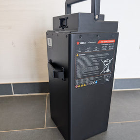 Ein schwarzer, rechteckiger Yadea-Lithium-LFP-Akku mit einem Griff steht auf einem Fliesenboden vor einer weißen Wand. An der Seite sind Warnaufkleber und technische Angaben zu sehen, die darauf hinweisen, dass er für den Einsatz im Yadea Owin U 2800W Retro-Elektroroller (45 km/h) geeignet ist.