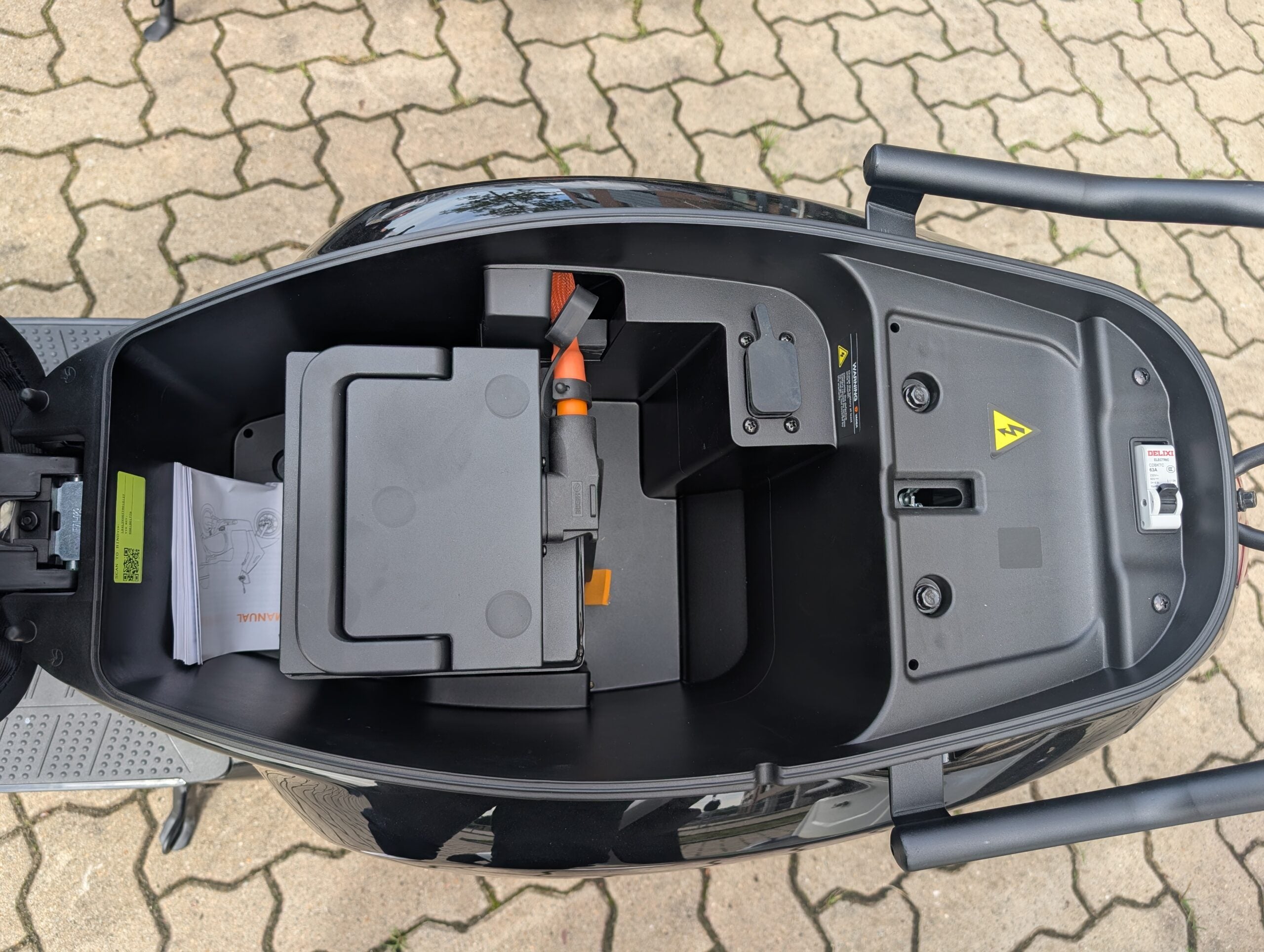 Öffnet man das Staufach unter dem Sitz des eleganten schwarzen Yadea Owin U 2800W Retro-Elektrorollers (45 km/h) von Yadea, kommen ein moderner LFP-Akku, einige Unterlagen und elektrische Bauteile zum Vorschein. Das Fach ist von einem schwarzen Rahmen umgeben und befindet sich auf gemustertem Pflaster.