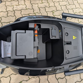 Öffnet man das Staufach unter dem Sitz des eleganten schwarzen Yadea Owin U 2800W Retro-Elektrorollers (45 km/h) von Yadea, kommen ein moderner LFP-Akku, einige Unterlagen und elektrische Bauteile zum Vorschein. Das Fach ist von einem schwarzen Rahmen umgeben und befindet sich auf gemustertem Pflaster.
