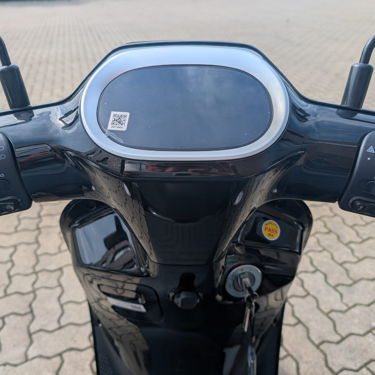 Nahaufnahme des Lenkers und des Armaturenbretts des schwarzen Yadea Owin U 2800W Retro-Elektrorollers (45 km/h) mit Bedientasten, einem Schlüssel im Zündschloss und einem auf dem Display sichtbaren QR-Code. Im Hintergrund ist eine gepflasterte Fläche zu sehen.